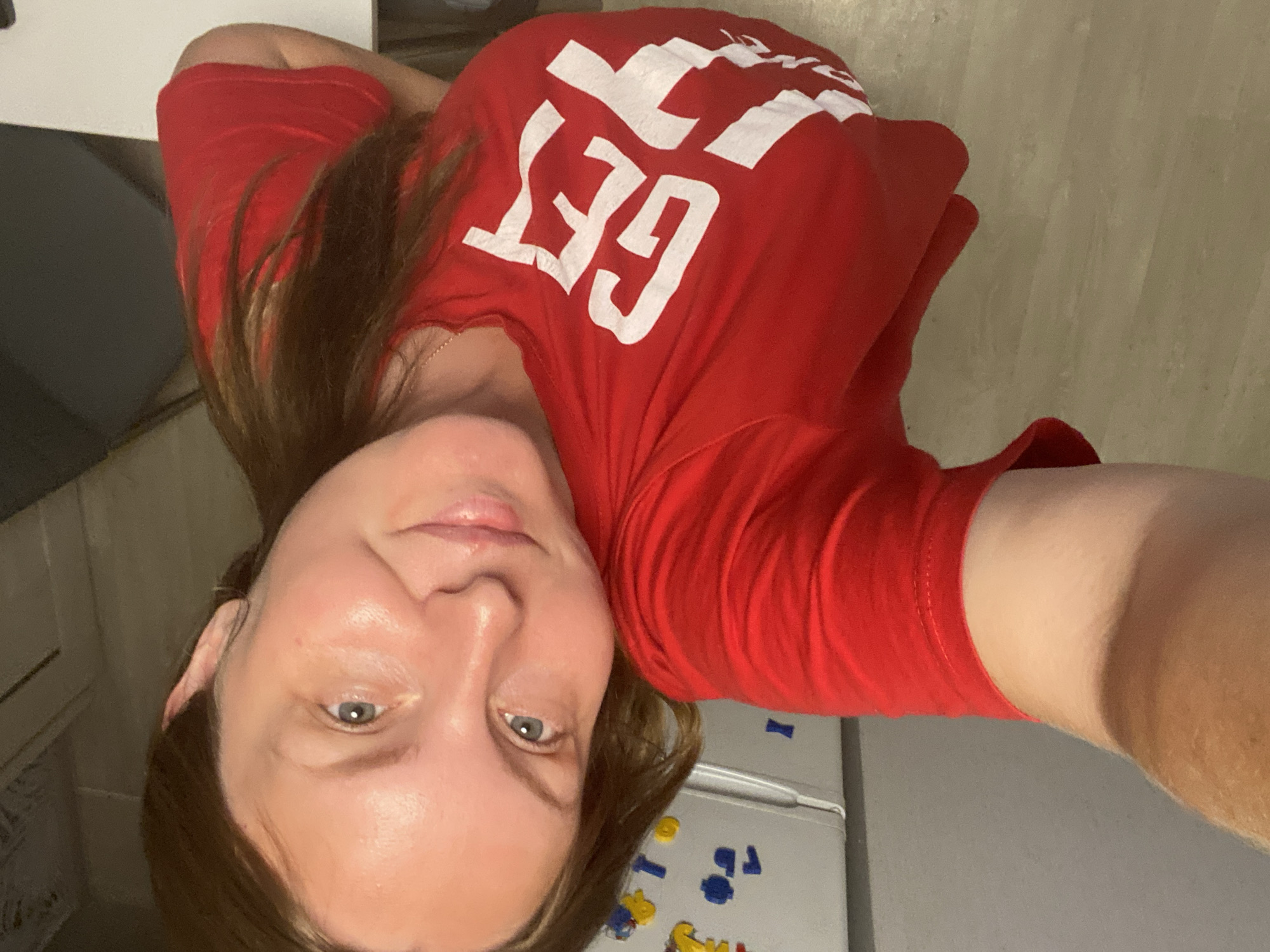 The image features a woman lying down, with her head resting on her arm. She is wearing a red t-shirt with the white text "GET IT" printed on it. The woman has light skin and brown hair that falls over her shoulder. Her eyes are open and she appears to be looking at the camera. The background suggests an indoor setting, with a neutral-colored wall and floor. There is also a hint of a refrigerator with colorful magnets in the bottom right corner of the image, suggesting a home environment. The lighting is dim, indicating it could be evening or an indoor setting with artificial light. The overall mood is casual and relaxed.