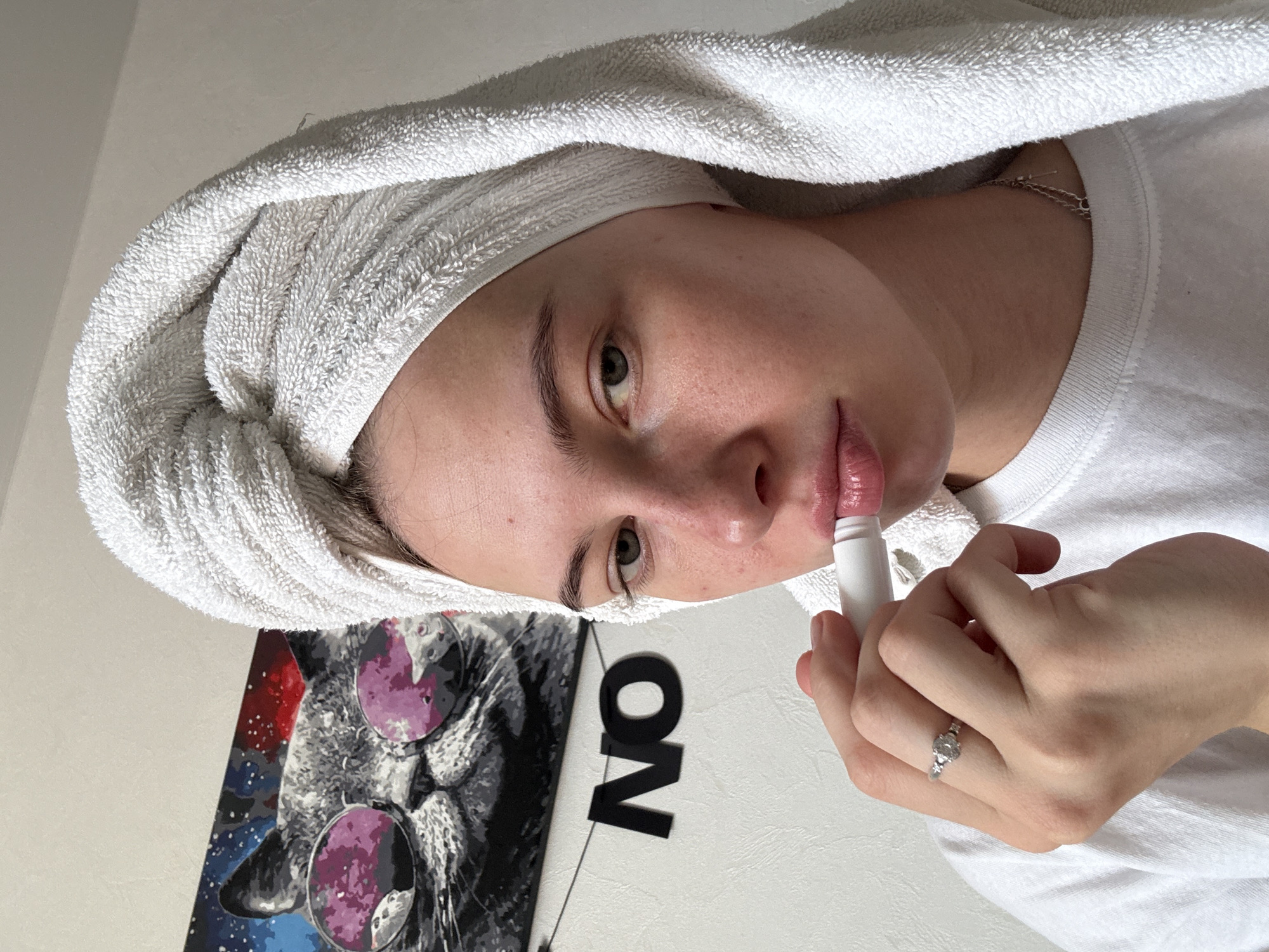A young woman with a white towel wrapped around her head is applying lip balm to her lips. She is wearing a white t-shirt and a silver ring on her left hand. The background features a painting of a cat wearing sunglasses and the word "NO" in black letters. The lighting suggests the image was taken indoors during the daytime. No specific location in Perm, Russia can be identified from the image.