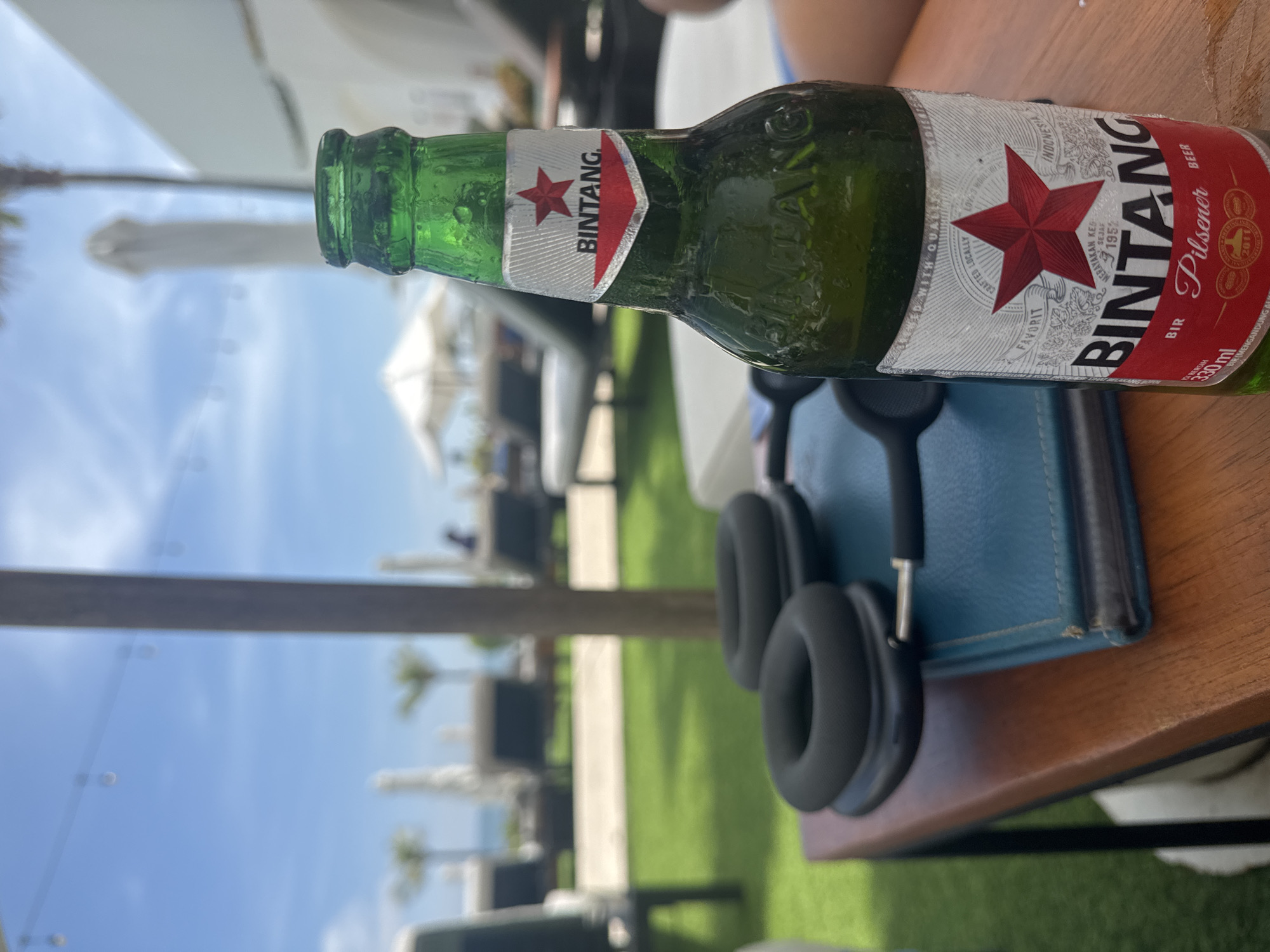 The image depicts a bottle of Bintang beer on a wooden table outdoors. The bottle is green and bears the Bintang logo, and appears chilled, as evidenced by condensation. Next to the bottle are a blue book and a pair of black headphones. In the background, the sky is visible, with the suggestion of an outdoor seating area under white umbrellas, indicating a likely tourist or resort setting. The location is implied to be North Kuta, Indonesia, based on the presence of Bintang beer, a popular local brand.