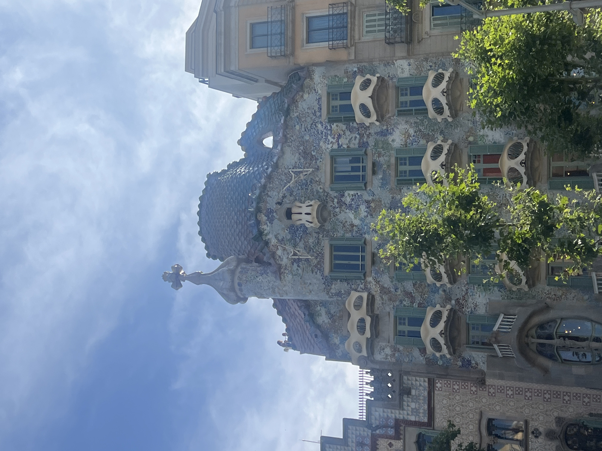 This is a vertical, eye-level photograph of Casa Batlló, a building designed by Antoni Gaudí, located on Passeig de Gràcia in Barcelona, Spain. The ornate facade is covered in colorful mosaic tiles and features unique architectural elements, including skeletal-like balconies and a dragon-shaped roof. The sky above is blue with scattered white clouds. Several trees with green foliage are visible in the foreground, partially obscuring the lower levels of the building. The building's distinctive roofline is topped with a turret and a cross. Some people are visible on the rooftop terrace of the building.