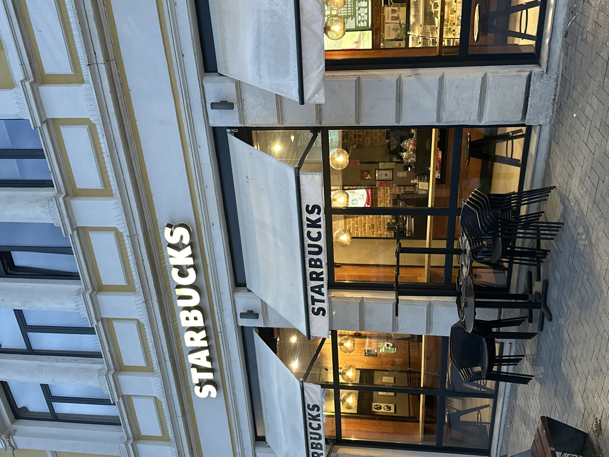 The image captures the exterior of a Starbucks coffee shop located on a street in Varna, Bulgaria. The scene appears to be set during twilight hours, either dusk or early morning, as the interior lights of the store are illuminated, casting a warm glow visible through the large glass windows, while the ambient light outside is dim.
The Starbucks occupies the ground floor of an ornate building with a light-colored facade featuring decorative architectural details and yellow-gold trim. Upper floors of the building contain dark-framed windows.
The coffee shop itself features a large, illuminated "STARBUCKS" sign prominently displayed on the building's facade. Below this, white retractable awnings are extended over the glass windows and entrance, each also bearing the "STARBUCKS" name in black lettering. Through the clear windows, several spherical light fixtures can be seen hanging inside, along with glimpses of the interior setup and brick wall sections.
On the sidewalk in front of the store, several black chairs are neatly stacked, and a few small tables are visible, indicating an outdoor seating area that is currently not in use, suggesting the store might be closed or preparing to open/close. The sidewalk is paved with grey bricks. The overall atmosphere is quiet and still.