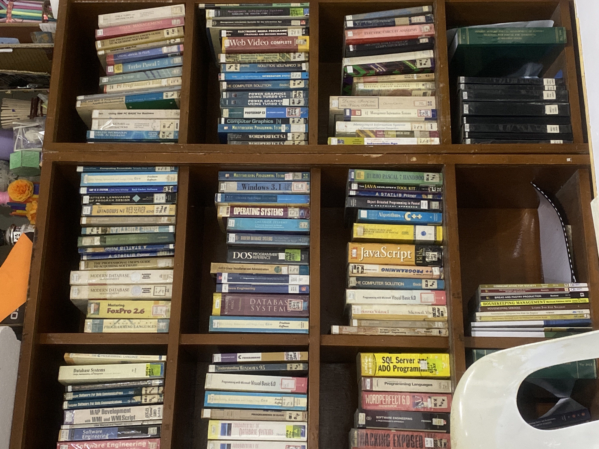 This media file presents a close-up, eye-level view of a multi-shelved wooden bookshelf, predominantly filled with older computer science and information technology books and software boxes. The setting appears to be an indoor space, possibly a personal study or a specialized library in Davao City, Philippines.
No people are visible, and no active event is taking place, depicting a static scene of stored items. The books are stacked vertically and horizontally, some appearing worn, indicating frequent use. A white chair is partially visible on the bottom right. On the left side, amidst the books, are a few decorative items, including an orange artificial flower and a green block, along with some loose papers.
The visible text on the book spines and software boxes reveals a strong focus on early programming languages, operating systems, and software development from the late 20th and early 21st centuries. Titles include "Turbo Pascal 7," "Windows 3.1," "DOS PROGRAMMER REFERENCE," "JavaScript Bible," "SQL Server," "Mastering FoxPro 2.6," "Operating Systems," "DATABASE SYSTEMS," "Software Engineering," "Computer Graphics," "PHOTOSHOP CS," "Web Video COMPLETE," "HACKING EXPOSED," and various "Management Information Systems" texts. Some outlier titles like "BREAD AND PASTRY PRODUCTION," "HOUSEKEEPING MANAGEMENT," and "COMMUNICATION FOR SOCIETY" are also present on the bottom right shelf, suggesting a broader range of interests or a mixed collection. The overall impression is a collection belonging to someone with a strong background or interest in older computing technologies.