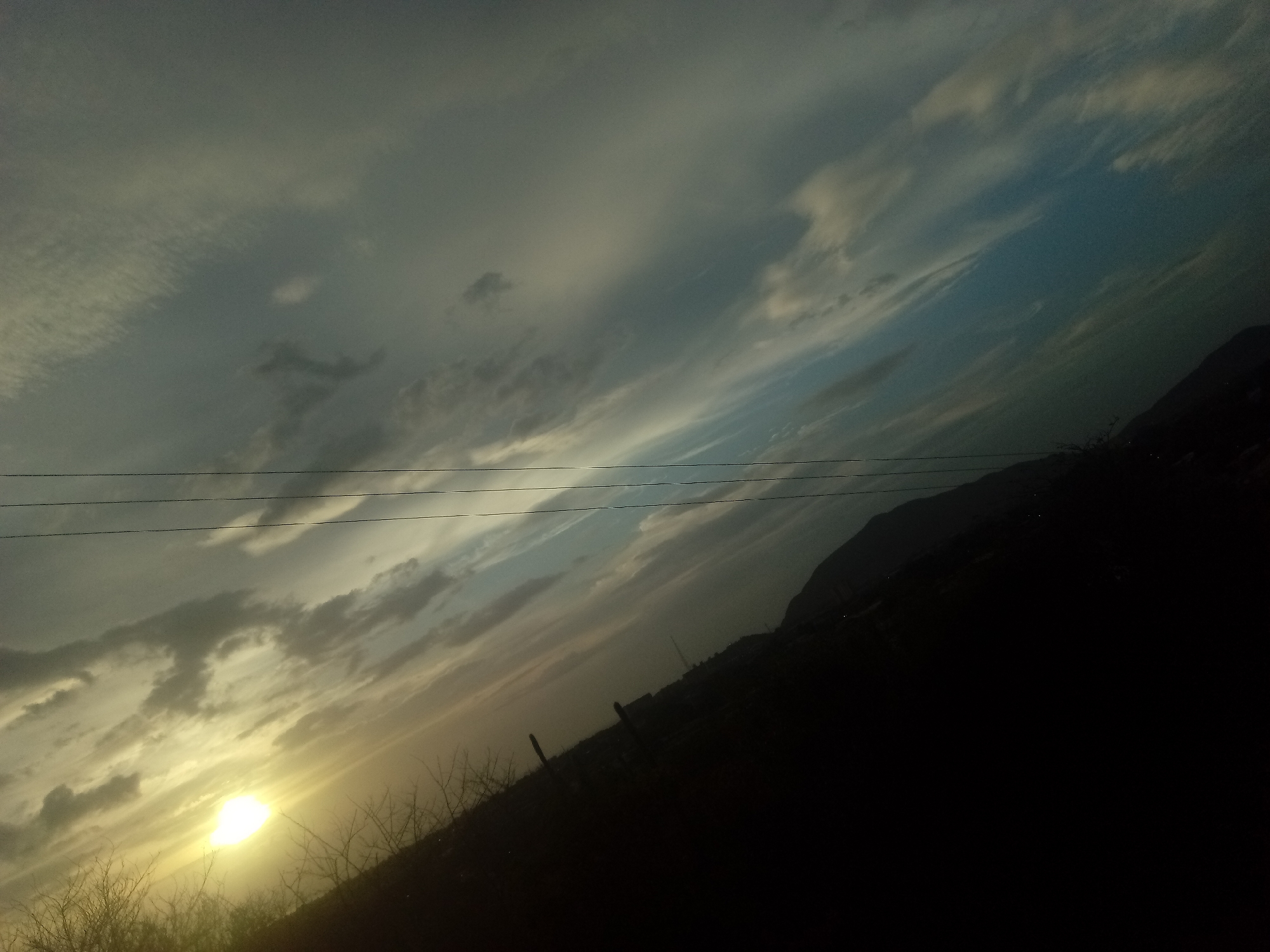 The image depicts a dusk scene in Altagracia, Venezuela. The sky is filled with dramatic clouds, illuminated by a setting sun visible on the lower left. Silhouetted telephone wires run across the upper part of the frame. A dark, sloped landmass occupies the lower right, likely a hillside or mountain, with faint indications of buildings or structures along its base. In the foreground, sparse vegetation is visible against the sunlit sky. The overall scene suggests a rural or semi-urban landscape in a mountainous area during twilight.