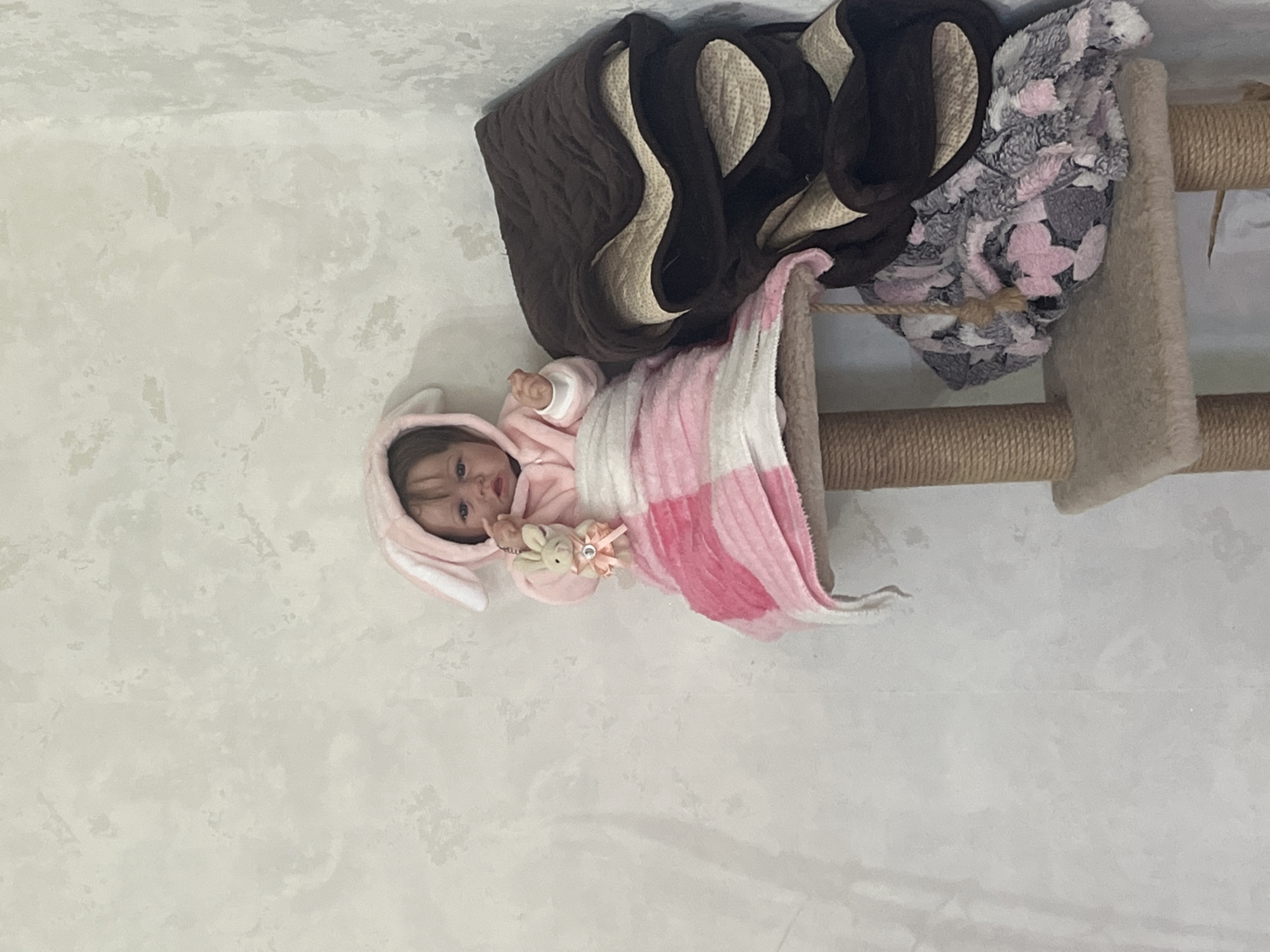 A doll with a pink bunny hood is sitting on a multi-tiered cat tree. The doll is wrapped in a pink and white checkered blanket and is holding a small plush bunny. The cat tree is located in a corner against a textured, light-colored wall. There are brown and gray patterned blankets stacked on the right side of the cat tree. The scene appears to be indoors in a domestic setting.
