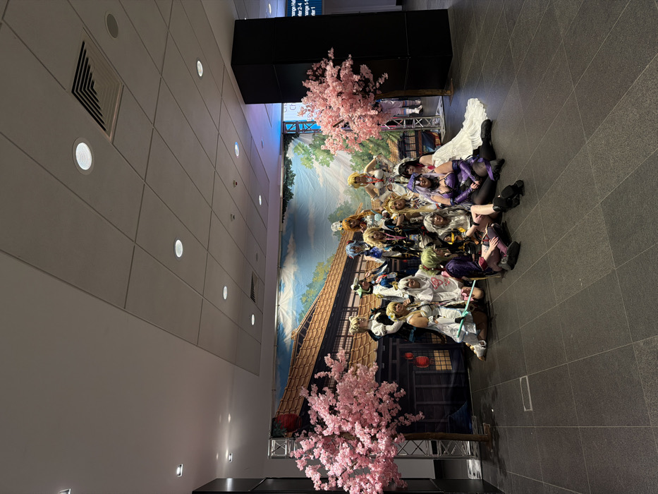 This image depicts an indoor group photograph of approximately 18-20 individuals, predominantly female, dressed in elaborate cosplay costumes, positioned within the Manchester Central Convention Complex in Manchester, United Kingdom.
The scene is set against a large fabric backdrop illustrating a traditional Japanese streetscape with wooden buildings, red lanterns, and a pale blue sky. Two artificial pink cherry blossom trees, featuring brown trunks and dense pink foliage, flank the group, positioned in front of the backdrop. Silver truss structures supporting the backdrop are partially visible on either side.
The individuals are arranged in a posed formation, with some standing, some kneeling, and others seated directly on the dark grey tiled floor. Most subjects are looking towards the camera. Their costumes exhibit diverse characters, featuring elaborate clothing, brightly colored wigs, and various accessories, including a person in a white costume holding a green-bladed prop sword.
The immediate environment consists of a dark grey tiled floor. The ceiling is a grid of white panels interspersed with numerous recessed circular lights and rectangular air vents. A prominent black rectangular pillar is visible on the right side of the frame, partially obscuring a white sign with black text that indicates "Orange Hall," "Trader 2-4," "Trader 1," and "Car Park 1," confirming the location within the Manchester Central Convention Complex. A dark, possibly black, vertical wall or pillar is present on the far left. The lighting appears even and artificial.
