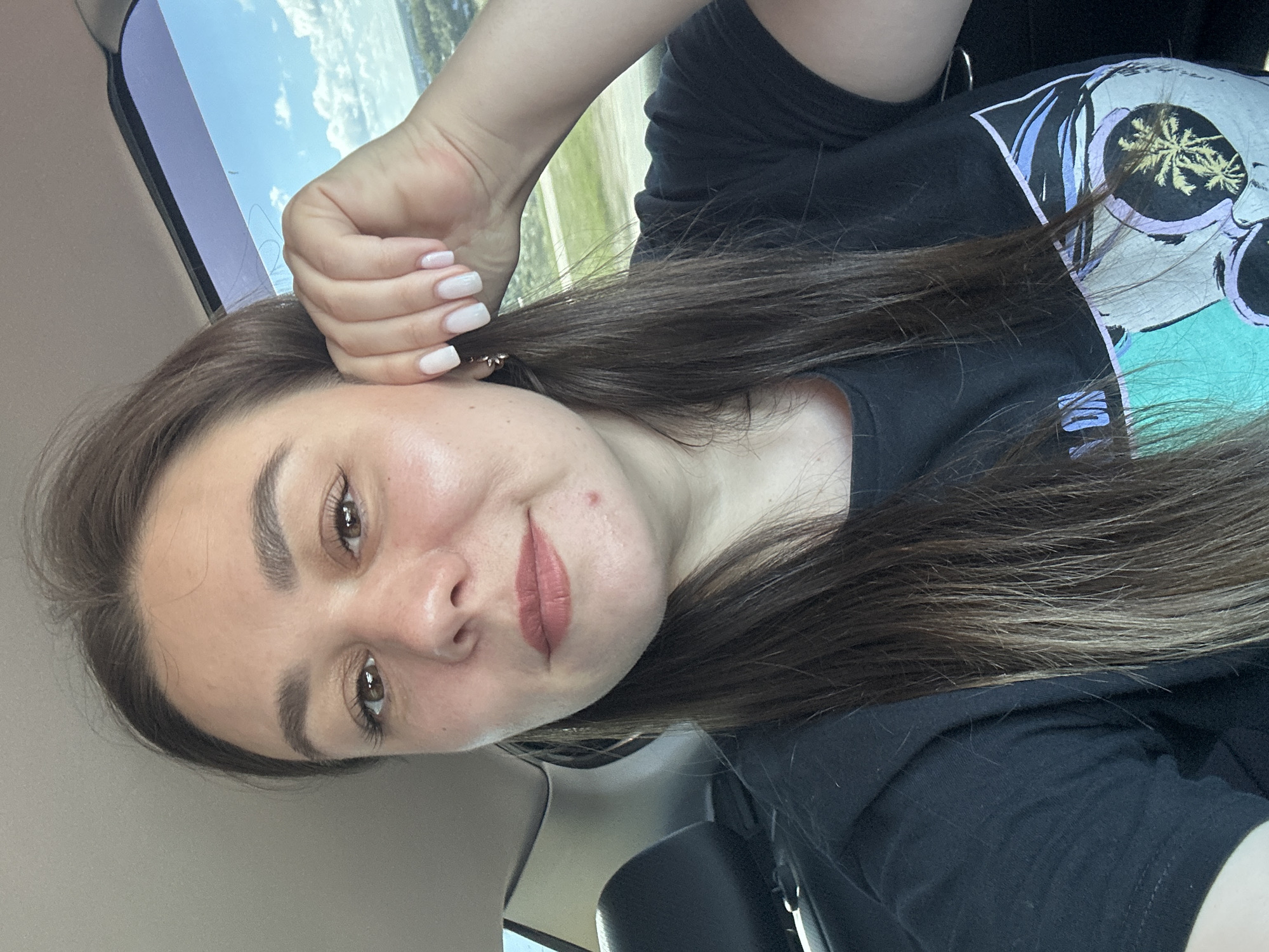 The image shows a close-up of a young woman with long, dark brown hair, wearing a black t-shirt. She is sitting in a car, with a window to her right showing a cloudy blue sky and a blurred landscape. Her right hand is raised to her ear, and her fingernails are painted white. She has a small blemish on her chin and is wearing light-colored lipstick. The background shows the interior of a car with a black seat visible on the left. The overall lighting suggests it is daytime. The provided location of Chaikovsky, Russia, is not discernible from the visual information in the image.