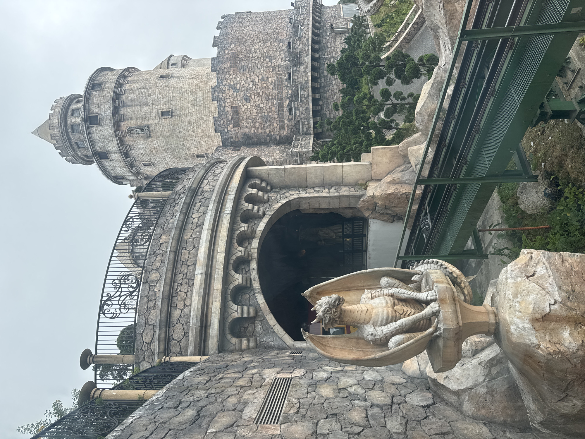 The image captures a fantasy-themed stone complex, resembling a grand castle or fortress, under an overcast sky. The primary structure is built from rugged stone blocks, featuring a prominent cylindrical tower with a pointed roof and smaller, intricately designed windows. Curving wrought-iron railings with decorative scrollwork adorn the balconies and staircases that spiral around parts of the tower.
In the foreground, a detailed stone statue of a winged dragon or gargoyle is mounted on a pedestal, its form conveying a sense of guardianship with its open mouth and outstretched wings. This statue is positioned near a large, dark arched entrance, framed by the textured stone architecture. The ground surrounding this area is paved with irregular stone and includes metal drainage grates.
To the right, a substantial green metal structure, likely part of a cable car or alpine coaster track with visible cables and support beams, extends across the scene. Beyond this, scattered trees and lush vegetation contribute to the outdoor setting. The overall weather appears cloudy or hazy, without direct sunlight, casting an atmospheric, muted light over the scene. No people or specific activities are depicted. The architectural style and prominent dragon motif are characteristic of fantasy-themed attractions found in places like Ba Na Hills, Đà Nẵng, Vietnam.