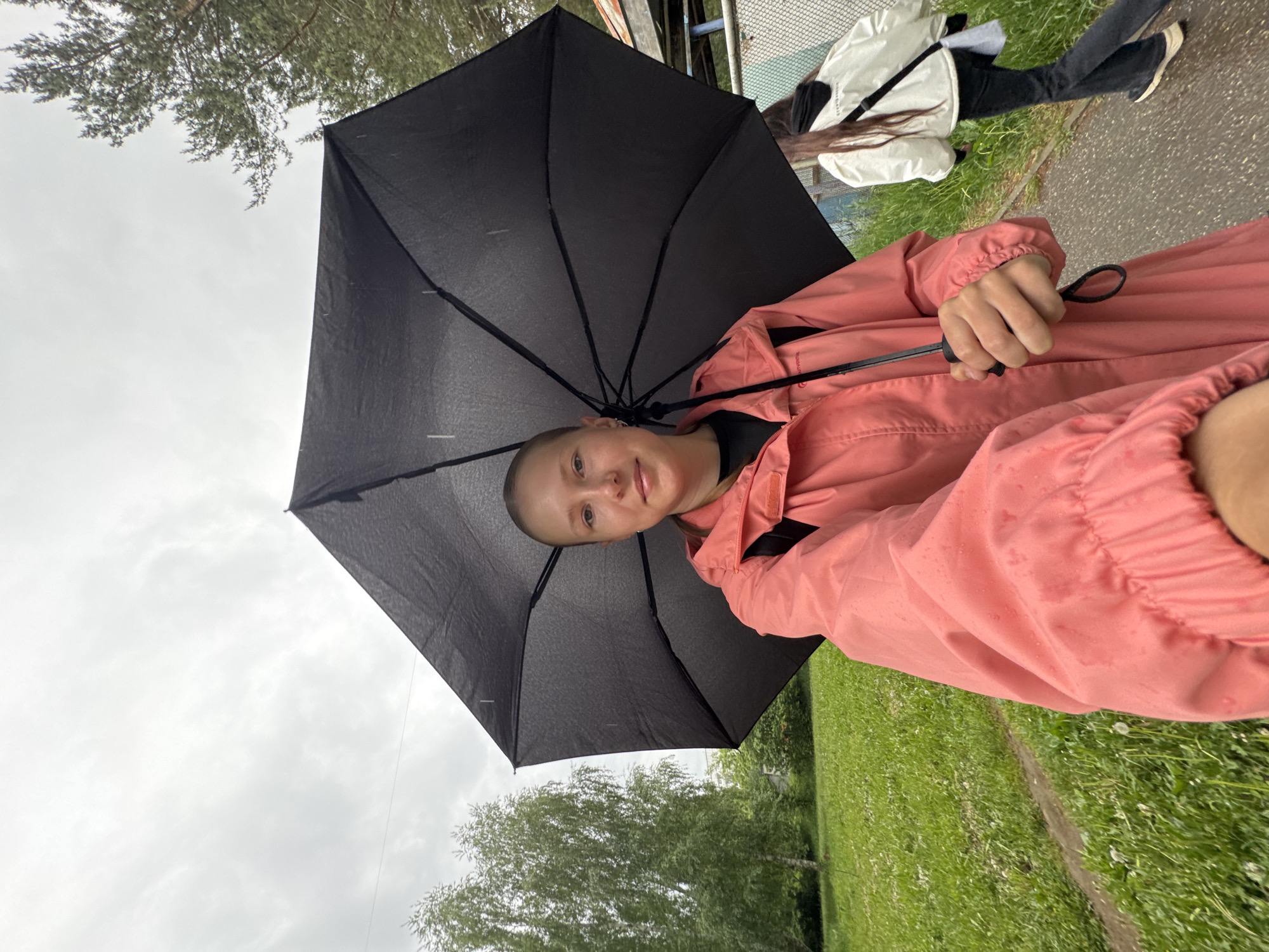 A young person with a shaved head and a friendly smile poses for the camera outdoors, holding a black umbrella. They are wearing a salmon-colored waterproof jacket over a black top. The jacket features a visible "OUTVENTURE ACTIVE LIFE" logo on the upper left arm. Raindrops are visible on the umbrella and the jacket, indicating it is raining or has recently rained.
In the background, another person walks away along a paved path, wearing a white jacket, black pants, and light-colored shoes, with long dark hair visible from under their jacket. The setting is an overcast day with diffused light. The ground is covered in vibrant green grass, and various trees are scattered around, including pine trees in the upper left and deciduous trees in the mid-ground. A metal fence or railing is partially visible in the upper right background. The scene depicts an everyday moment in Chaikovsky, Russia, likely during inclement weather.