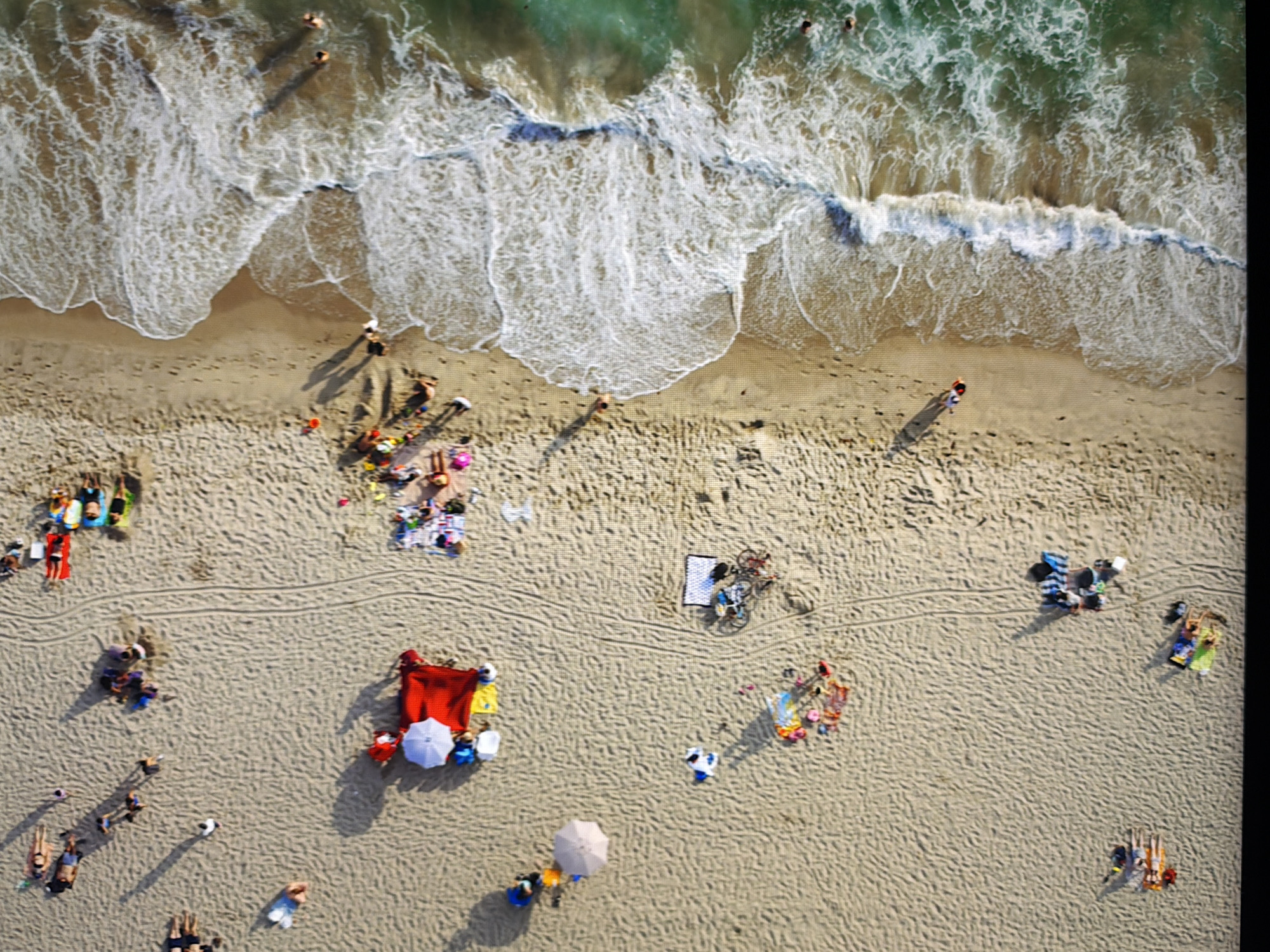 This is an aerial, high-angle, full shot of a sandy beach with a body of water. There are numerous people on the beach, some sunbathing on towels, others walking or standing. Several beach umbrellas are visible, providing shade. A red blanket is spread out with people sitting around it. There are also a couple of bicycles parked on the sand. The water is a turquoise green with white foamy waves reaching the shore. The scene depicts a typical beach day with various recreational activities. The image is not taken in Kyiv, Ukraine.