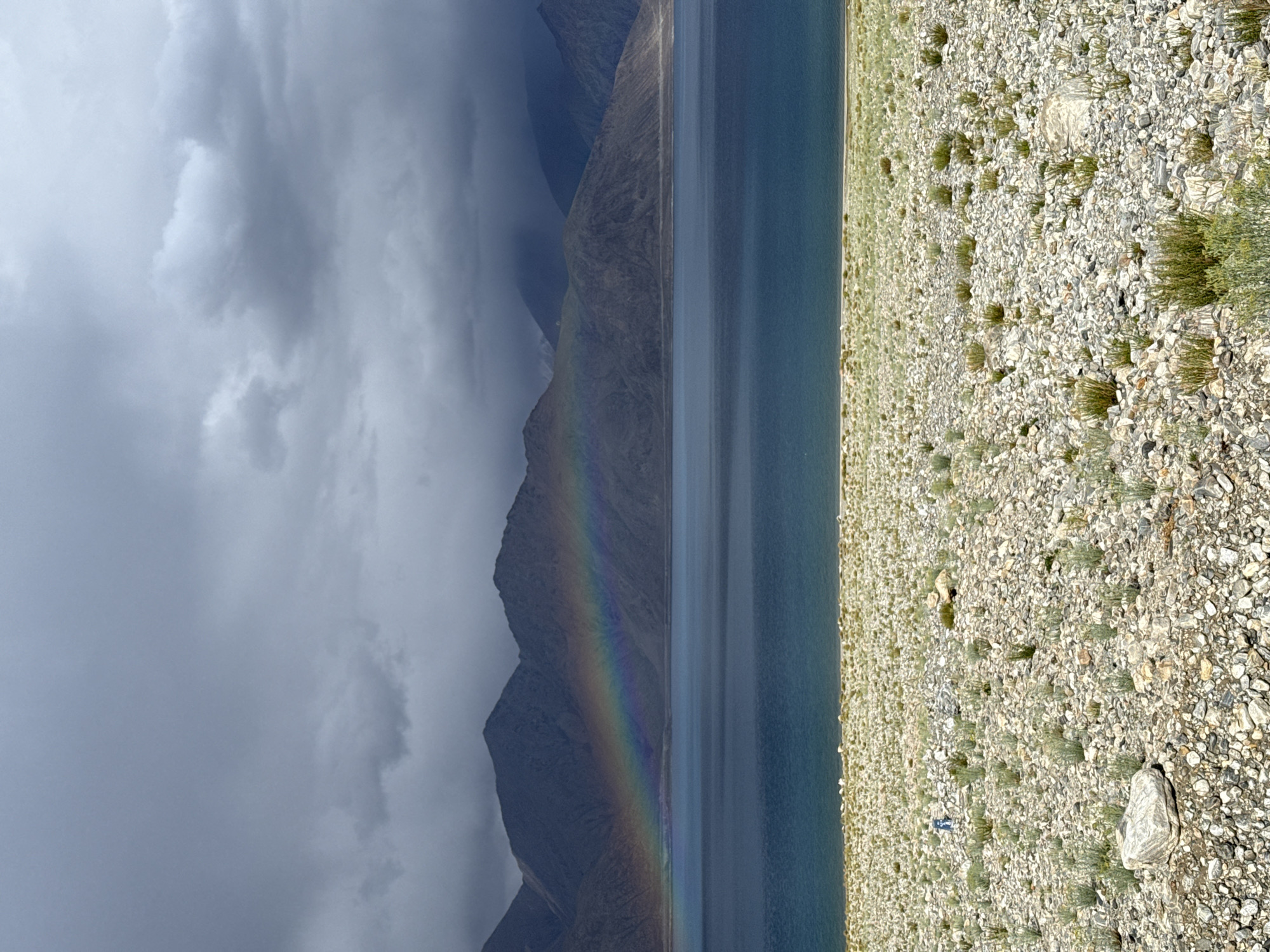 The image depicts a panoramic view of Pangong Tso lake in Man, India. The lake is vast and tranquil, with deep blue water reflecting the stormy sky. A vibrant rainbow arcs across the sky, a stark contrast to the dark, moody clouds that dominate the upper portion of the frame. Jagged mountains form a dramatic backdrop to the scene, their slopes appearing barren and rocky. In the foreground, the shore is covered in a dense layer of pebbles and sparse, dry vegetation. A solitary figure, appearing as a small silhouette, walks along the shoreline, adding a sense of scale to the immensity of the landscape. The overall impression is one of raw, untamed natural beauty, with a sense of both awe and foreboding.
