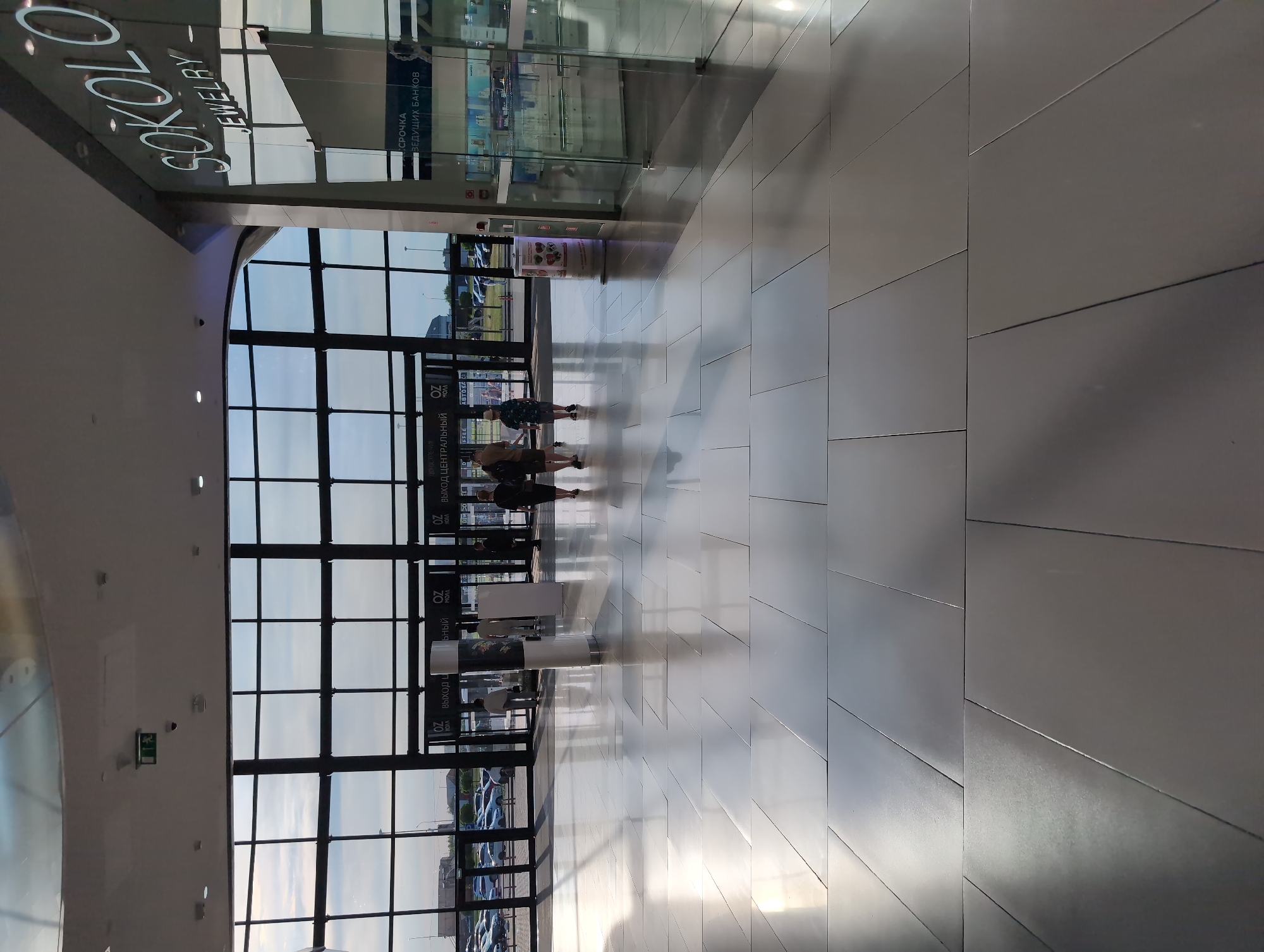 The image depicts the interior of a modern shopping mall in Krasnodar, Russia, specifically at the OZ Mall. In the foreground, a large, polished, light-colored tiled floor reflects the ambient light, creating a bright and spacious atmosphere. Several large, floor-to-ceiling windows dominate the background, offering a view of an outdoor area with cars and a cloudy sky. Through the windows, one can discern the entrances and exits of the mall, marked with signage.
To the right of the image, a prominent store facade displays the "SOKOLO JEWELRY" logo in large, white, illuminated letters. Adjacent to the jewelry store is a glass-enclosed space, possibly another shop or service area.
Several individuals are visible in the mid-ground, walking across the floor. One group of three people, two women and a man, are walking towards the right side of the frame. Another person, a man, is walking towards the left. The individuals are casually dressed.
In the middle ground, a cylindrical display stand with advertisements or information is positioned. A large white rectangular stand is also present nearby. The overall environment suggests a busy commercial space designed with modern aesthetics, emphasizing light, space, and polished surfaces. The lighting appears to be a combination of natural light from the windows and artificial lighting from the ceiling.