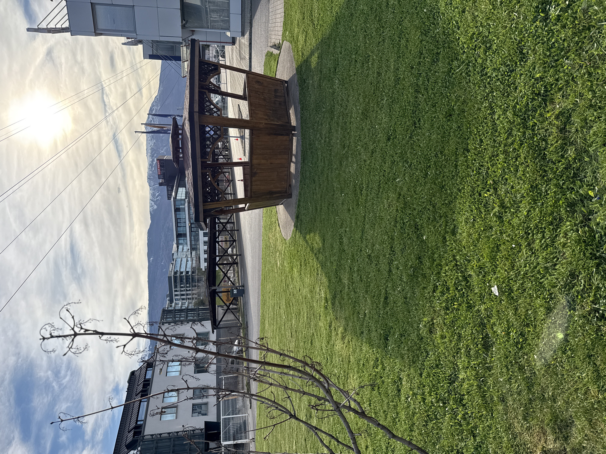 The image shows a grassy area next to a modern building with a small wooden structure. A tree with bare branches is in the foreground. Mountains are visible in the background with a blue sky and the sun is shining on a cloudy day. There is a cable car passing over a building. The location is unknown and could not be determined.