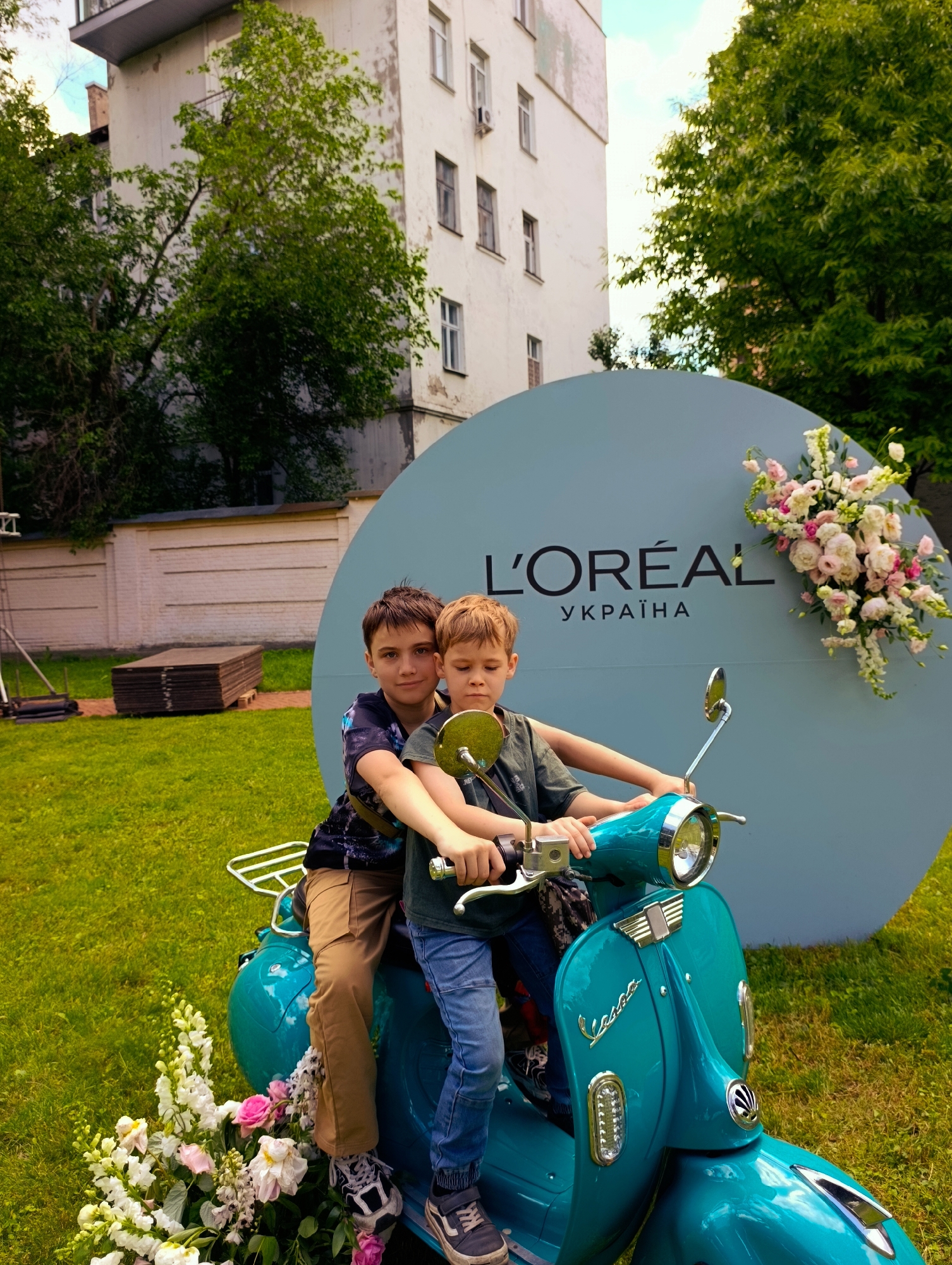 Two boys are posing on a teal Vespa scooter in front of a large, circular light blue backdrop featuring the L'Oréal logo and text in Ukrainian. The backdrop is decorated with a floral arrangement of pink and white flowers on the right side. The scooter is positioned on a well-maintained grassy area. In the background, a white building with multiple windows and balconies is visible, partially obscured by green trees. To the left of the building, a brick wall and stacked wooden planks are present. The scene appears to be an outdoor event or promotional setup, possibly in Kyiv, Ukraine, given the Ukrainian text on the backdrop. The older boy is seated behind the younger boy, who is in the driver's seat. Both boys are looking towards the camera.