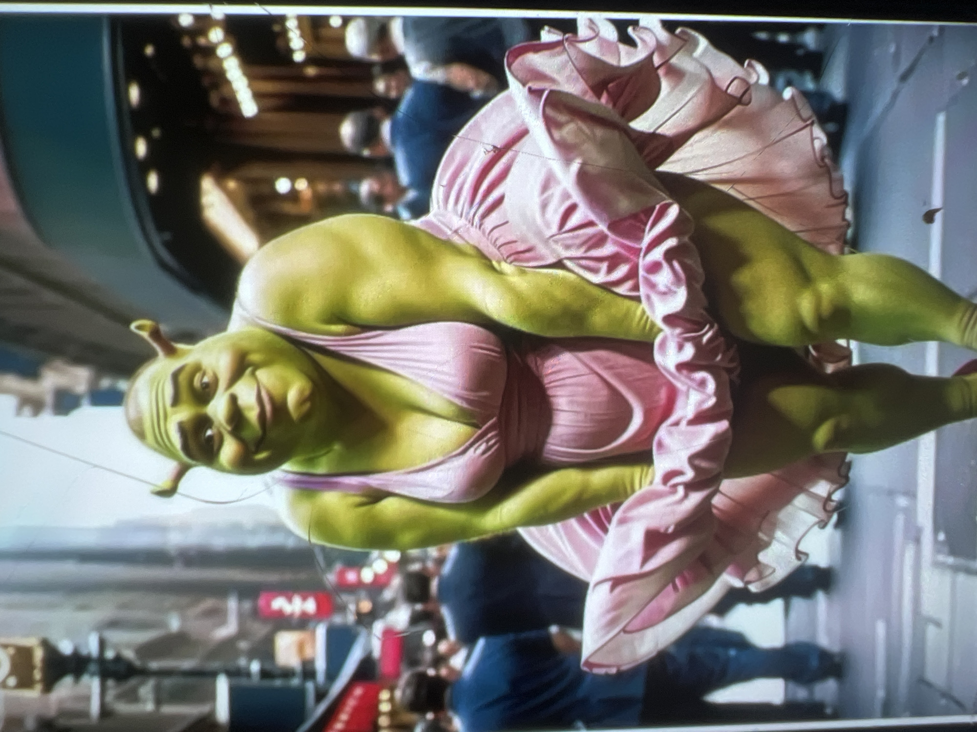 The image shows a depiction of Shrek from the animated film franchise, rendered with a human-like physique and wearing a pink Marilyn Monroe-esque dress, positioned in a city street. Shrek's skin is its characteristic green hue, and the dress is a bright pink satin with ruffles. He is smiling broadly and looking towards the viewer. The background appears to be a busy city street, possibly New York City, with buildings and blurred figures of people in the distance. There are no elements in the image that suggest the location of Kara-Dıykan, Kyrgyzstan.