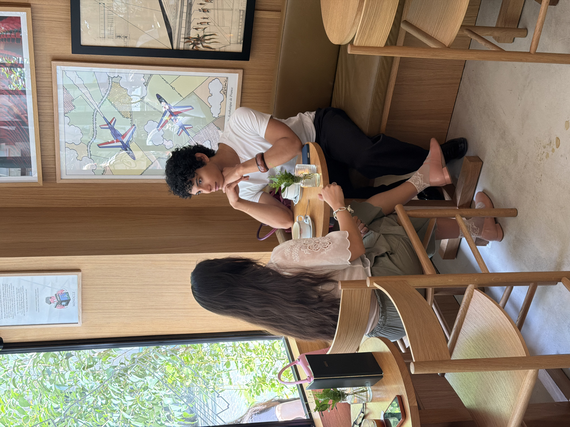 Two people are seated at a small round table in what appears to be a café or restaurant. The person on the left has long, dark hair and is wearing a light-colored blouse. A black notebook and eyeglasses are visible on the table. The person on the right has short, curly dark hair and is wearing a white T-shirt and black pants. The setting includes wooden chairs and tables, artwork on the walls depicting planes, and a framed advertisement for MONOCLE magazine. Natural light streams in from a window on the left side. The atmosphere appears relaxed and intimate. It is not possible to confirm if this is in Paris, France.