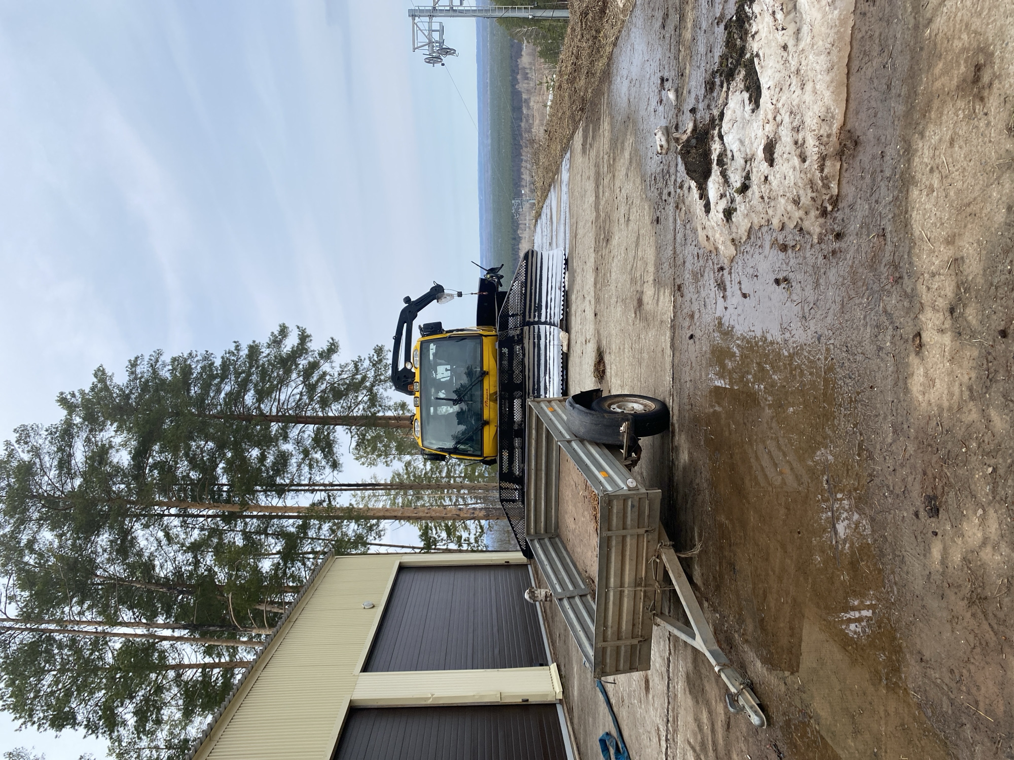 The image depicts a yellow snow groomer with a trailer parked on a muddy, concrete surface in Прикамский, Russia. Behind the groomer are tall pine trees and a building with closed brown garage doors. In the background, there is a ski lift and a forest in the distance. Patches of snow are visible on the ground, indicating a cold climate. The sky is pale blue, suggesting an overcast day.