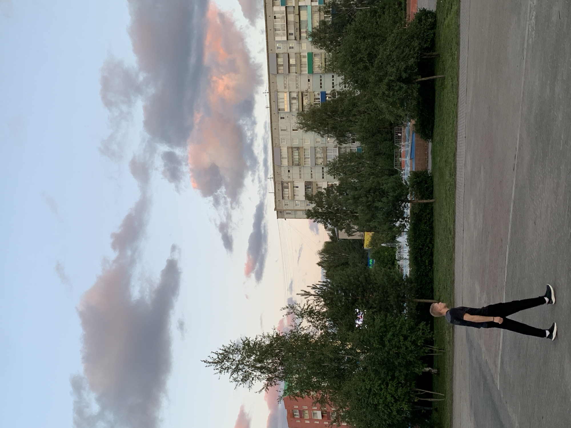 The image depicts a street scene in the city of Prokopyevsk, Russia, during a soft twilight. A young Caucasian man with blond hair walks from left to right across a paved walkway. He is dressed in a black t-shirt and black pants, with white-soled black sneakers. His posture suggests a casual stroll. Behind him, a strip of green grass leads to a line of trees and bushes. In the background, a large, light-colored apartment building with numerous windows stretches across the frame. To the left of the apartment building, a portion of a red-brick building is visible. The sky above is a gradient of pale blue, transitioning to pink and gray clouds, suggesting either sunrise or sunset. The overall atmosphere is calm and urban.