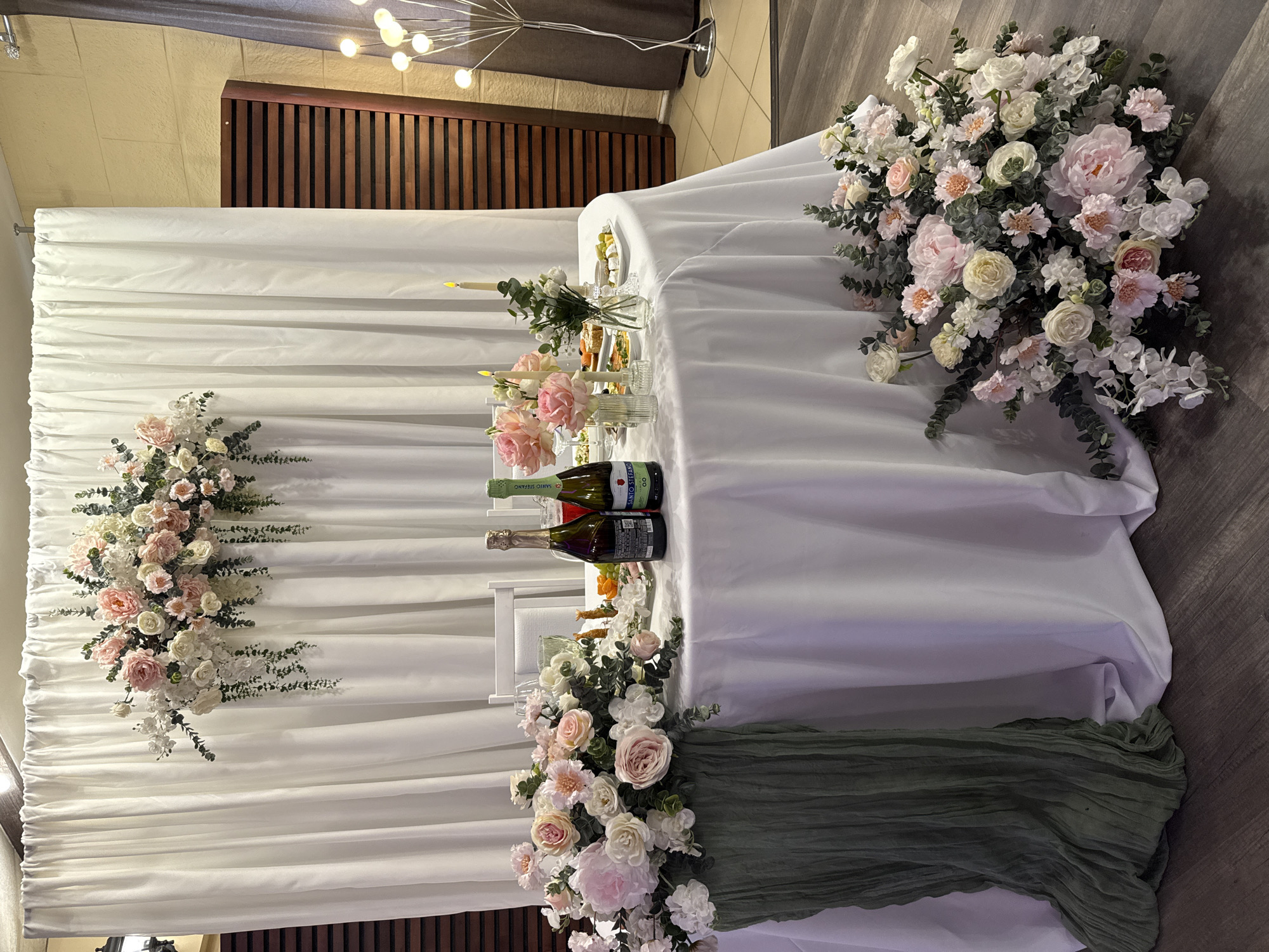 The image depicts a wedding reception setup. A long white table is draped with fabric, and floral arrangements are placed on the table and against the white backdrop behind it. Bottles of what appear to be wine or champagne are visible on the table. Cutlery and glasses suggest that food will be served at the table. A green cloth is visible at the bottom right of the table. The specific location is not discernible from the image. The image contains no people. The city of Chaikovsky is not discernible.