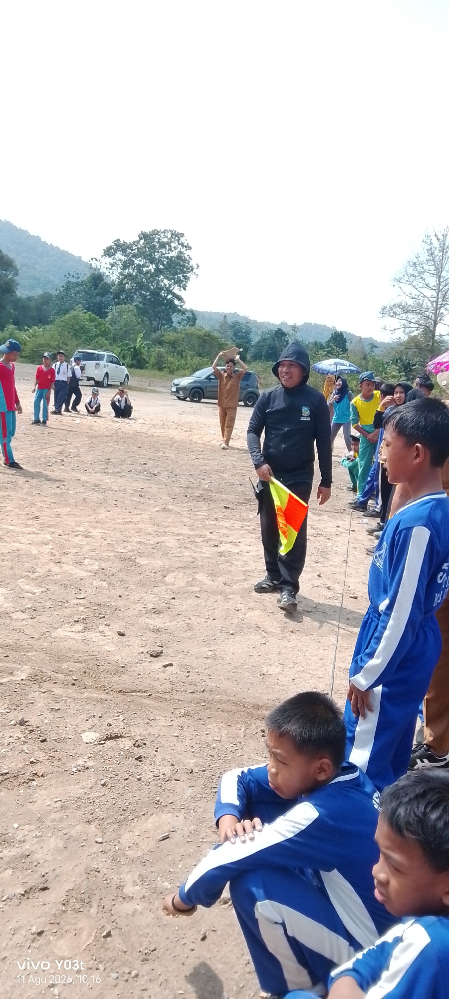 In Tirawuta, Indonesia, a group of children and adults gather on a dirt field. In the foreground, two young boys in blue tracksuits are crouched down, looking to the right. A man in a black hooded jacket and black pants stands in the center, holding a yellow and orange checkered flag. He appears to be a referee or official. Behind him, other people are visible, including some children in school uniforms and a man carrying a flat object on his head. Two cars are parked in the background, with a lush, green hillside behind them. The sky is bright and clear, suggesting a sunny day.