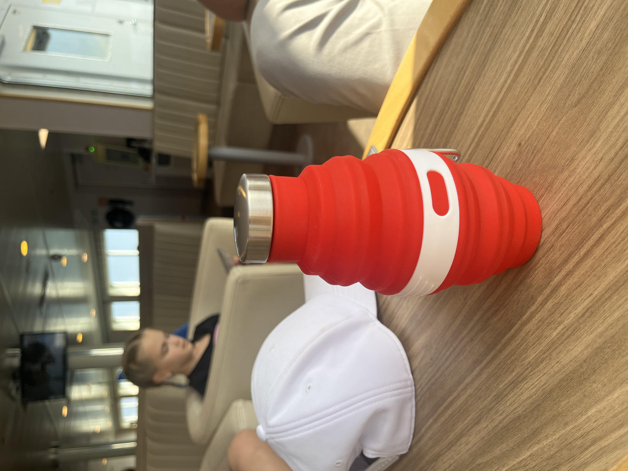 A red, collapsible water bottle with a white band and a silver cap sits on a wooden table. Next to the bottle is a white baseball cap. In the background, a young girl with braided blonde hair is seated in a beige booth, looking down at a book or device. The scene appears to be indoors, possibly on a ferry, given the windows and seating arrangement. The location is identified as Virtsu, Estonia.