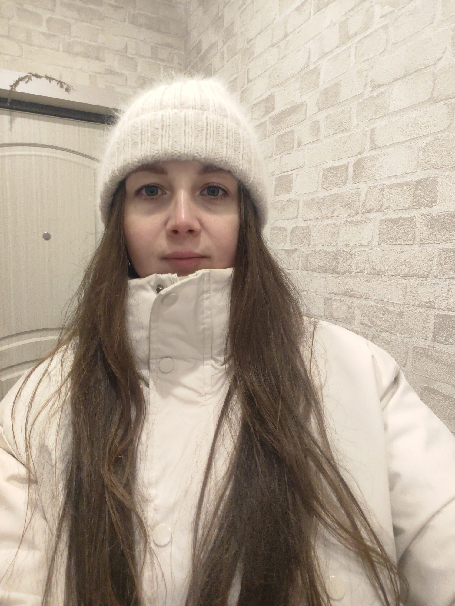 A waist-up indoor shot of a woman wearing a white knitted hat and a white puffer jacket. She has long brown hair. The background is a light-colored brick wall and a closed light-colored door. Location: Unknown, Unknown.