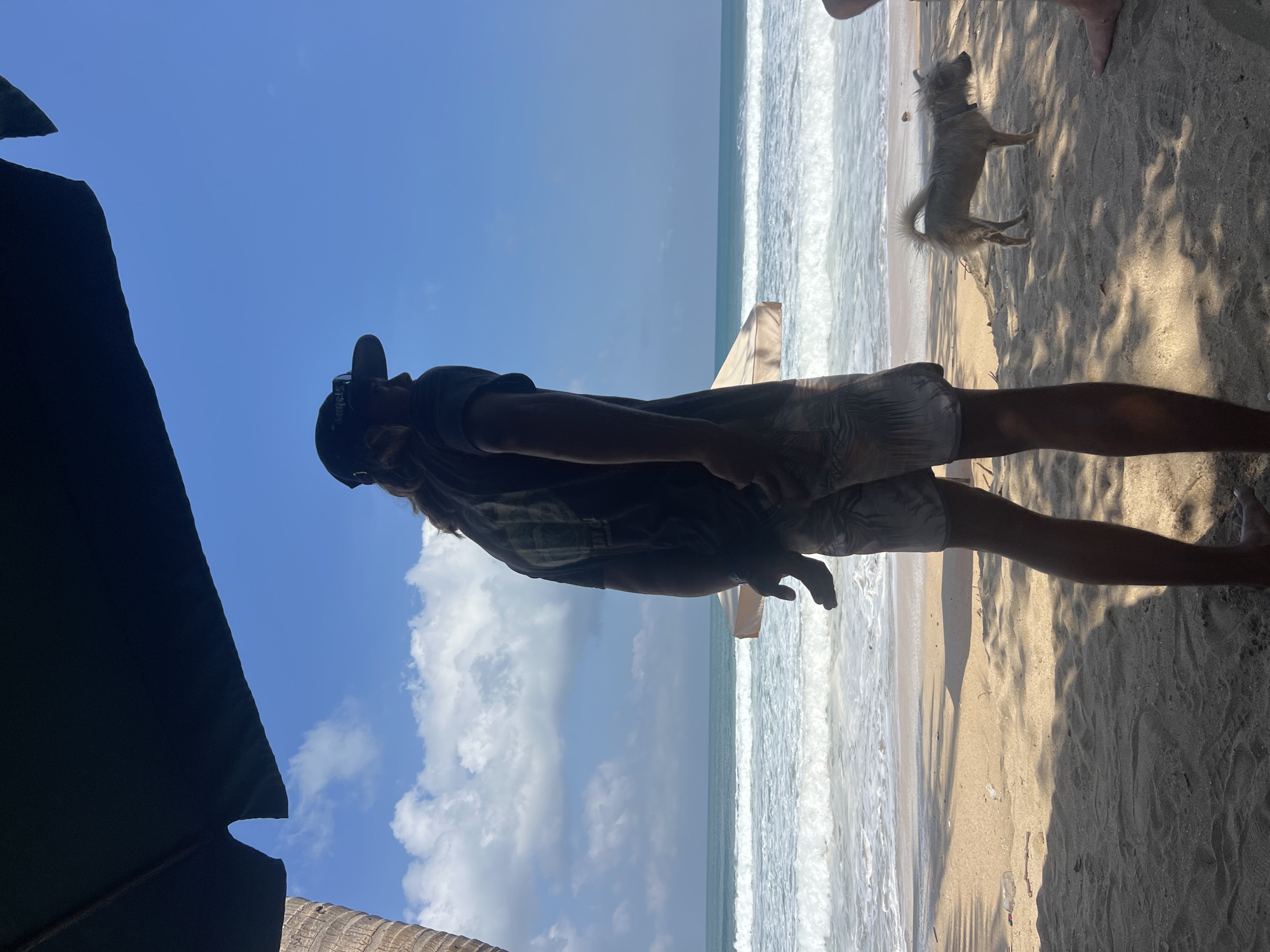 A man wearing a black t-shirt, patterned shorts, a dark baseball cap, and sunglasses is standing on a sandy beach, facing away from the camera and looking out at the ocean. He is partially shaded by a large dark umbrella or canopy and a palm tree trunk is visible to his left. In the background, the ocean has moderate waves and the sky is blue with scattered clouds. A small, light-colored dog stands on the sand to the man's right, also looking towards the ocean. Another person's bare feet are visible at the far right of the frame. The location is Kuta Beach, Bali, Indonesia, indicated by the distinctive palm trees, surf conditions, and the overall atmosphere of a popular tourist beach.
