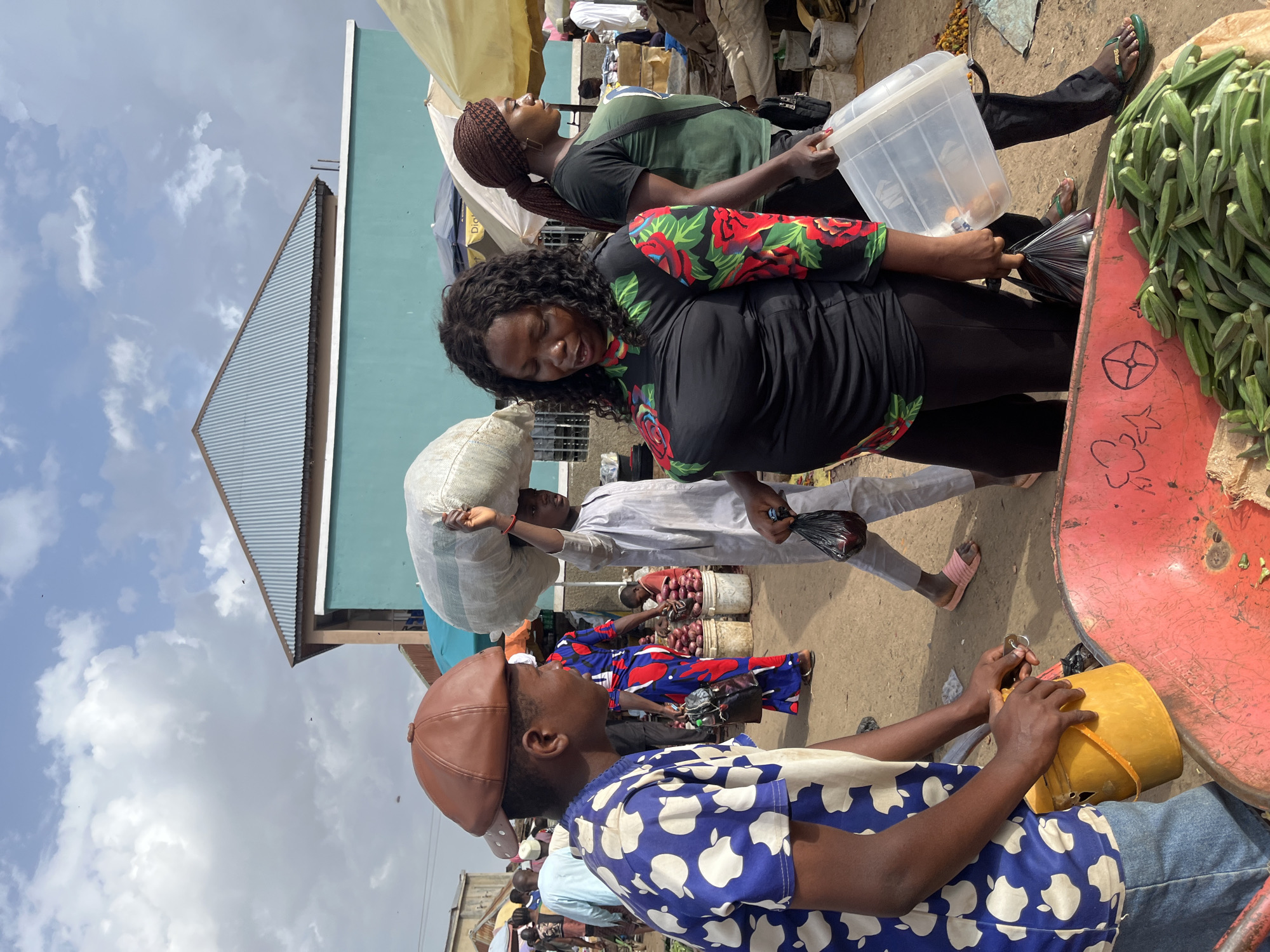 In Rafin Par, Nigeria, a bustling market scene unfolds under a partly cloudy sky. A person wearing a blue and white patterned shirt and a brown cap is seen holding a yellow bucket, facing a woman in a black shirt with a floral design. The woman is looking down, seemingly engaged in conversation, and is holding a black plastic bag and a clear plastic container. Behind them, another individual in a gray traditional outfit carries a large sack of goods on their head. The market appears to be an outdoor setting with various stalls and produce, including a wheelbarrow filled with okra in the foreground. The ground is a dusty, earthy surface.