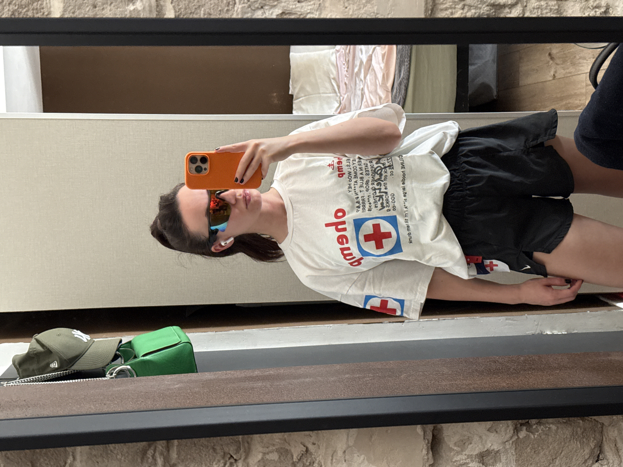 A young woman is taking a selfie in front of a mirror. She is wearing sunglasses, black shorts, and a white t-shirt with a red cross graphic on it. She holds an orange-colored smartphone. A green purse and a green baseball cap are visible on the ledge of the mirror. The location is implied to be Tel-Aviv, Israel. The background is blurred, showing a room interior.