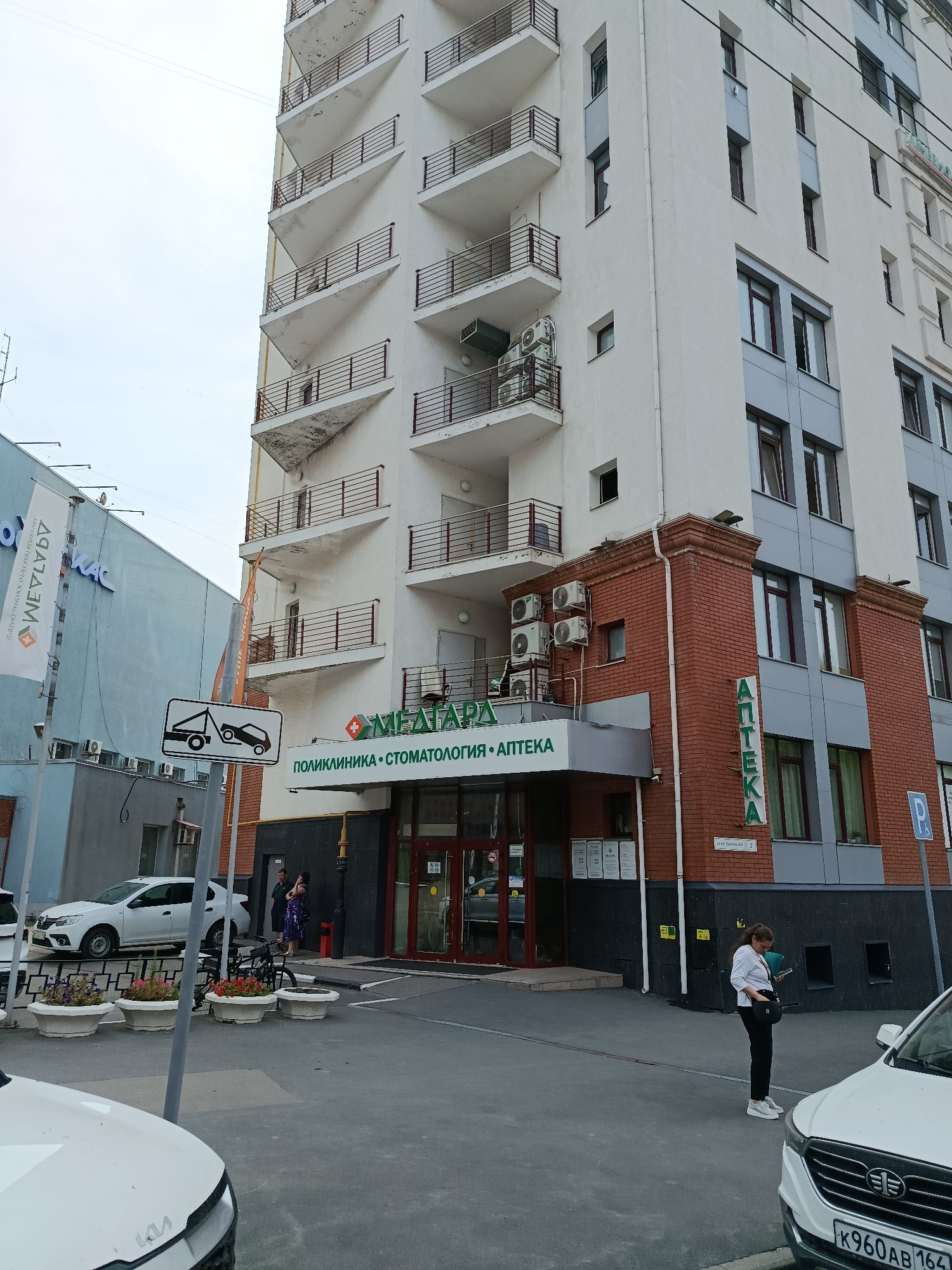 A multi-story building in Saratov, Russia, features a brick lower section and a white upper section with multiple balconies. A sign above the entrance reads "МЕДАРТА" (MEDARTA) followed by "ПОЛИКЛИНИКА • СТОМАТОЛОГИЯ • АПТЕКА" (POLYCLINIC • DENTISTRY • PHARMACY). To the right, a vertical sign spells out "АПТЕКА" (APTEKA). Several air conditioning units are mounted on the building's exterior. A tow-away zone sign is visible on a pole in front of the building. A white sedan is parked on the street, and a white car is partially visible in the foreground. A woman in dark clothing is standing near the entrance, and another woman in white and black is looking at her phone near a parked car. Planters with flowers are arranged along the sidewalk. The sky is overcast.