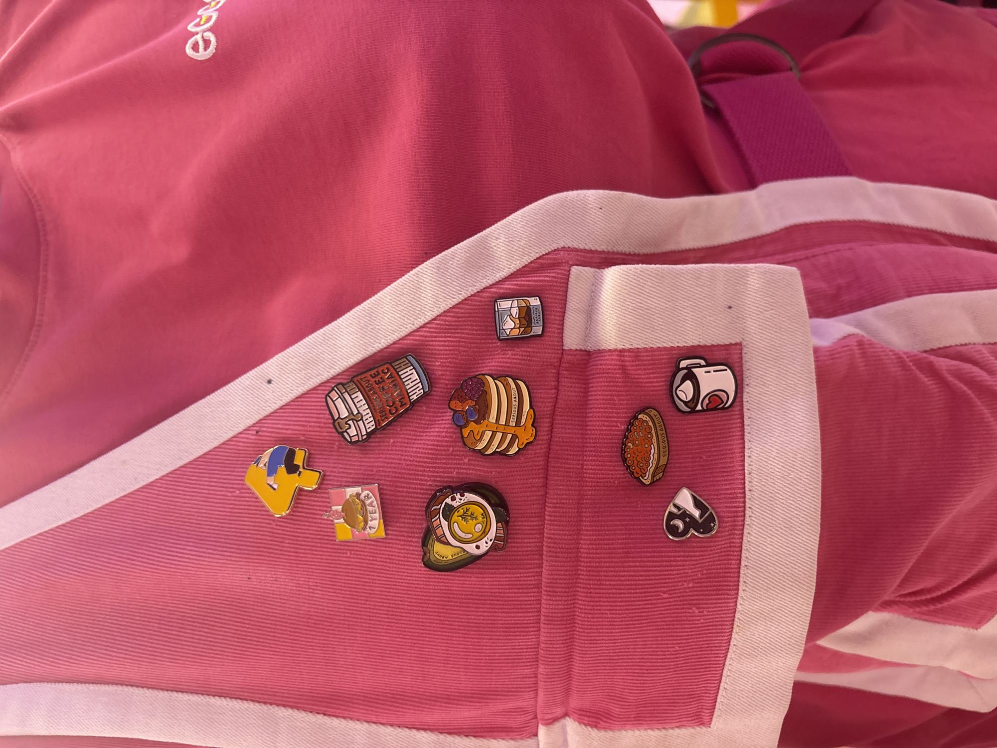 The image shows a section of a pink garment, possibly a bag or jacket, adorned with various enamel pins. The garment has white trim and a corduroy texture. The pins depict a variety of food items including pancakes, coffee, avocado toast, and other breakfast-themed designs. Some pins have text on them, such as "Serious About" and "Coffee About it." The overall setting is a close-up view with no background visible, and there is no evidence to suggest the image was taken in Moscow, Russia. There are no people in the image.