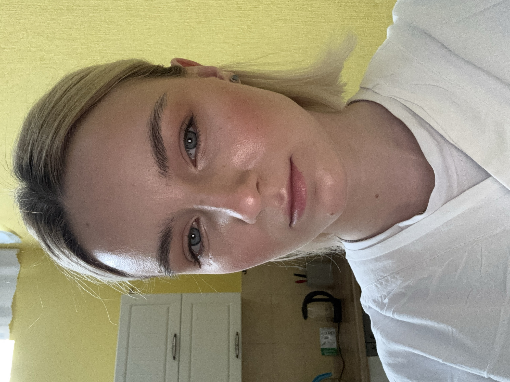 A close-up, slightly rotated image features a fair-skinned woman with short, light blonde hair. She has blue eyes and defined eyebrows. Her skin appears to have a subtle blush tone. She's wearing a white shirt with visible creases. The background is yellow in the upper portion of the image, transitioning to a kitchen-like setting with white cabinets and a dark object that might be a headphone. The location is indeterminable from the image.