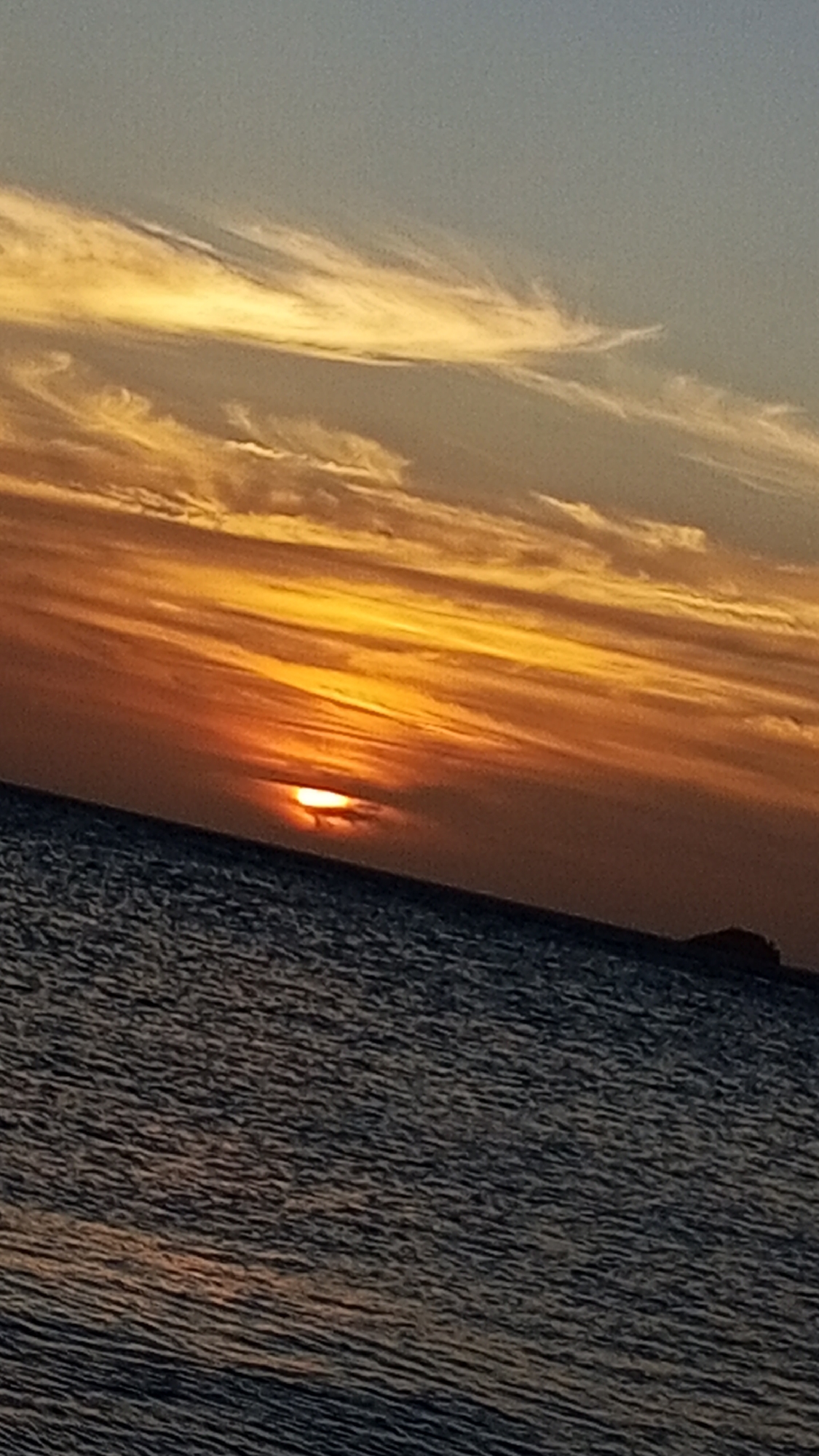 The image depicts a sunset over water, likely the Caribbean Sea, near Pedro González, Venezuela. The sun is partially obscured by a layer of clouds near the horizon, casting a bright orange and yellow glow across the sky. Streaky, wispy clouds are present in the upper portion of the sky. The water appears dark and textured with ripples. A small, dark object, possibly an island or distant landmass, is visible on the horizon towards the right side of the frame. No people are visible. The overall scene conveys a sense of tranquility and natural beauty.