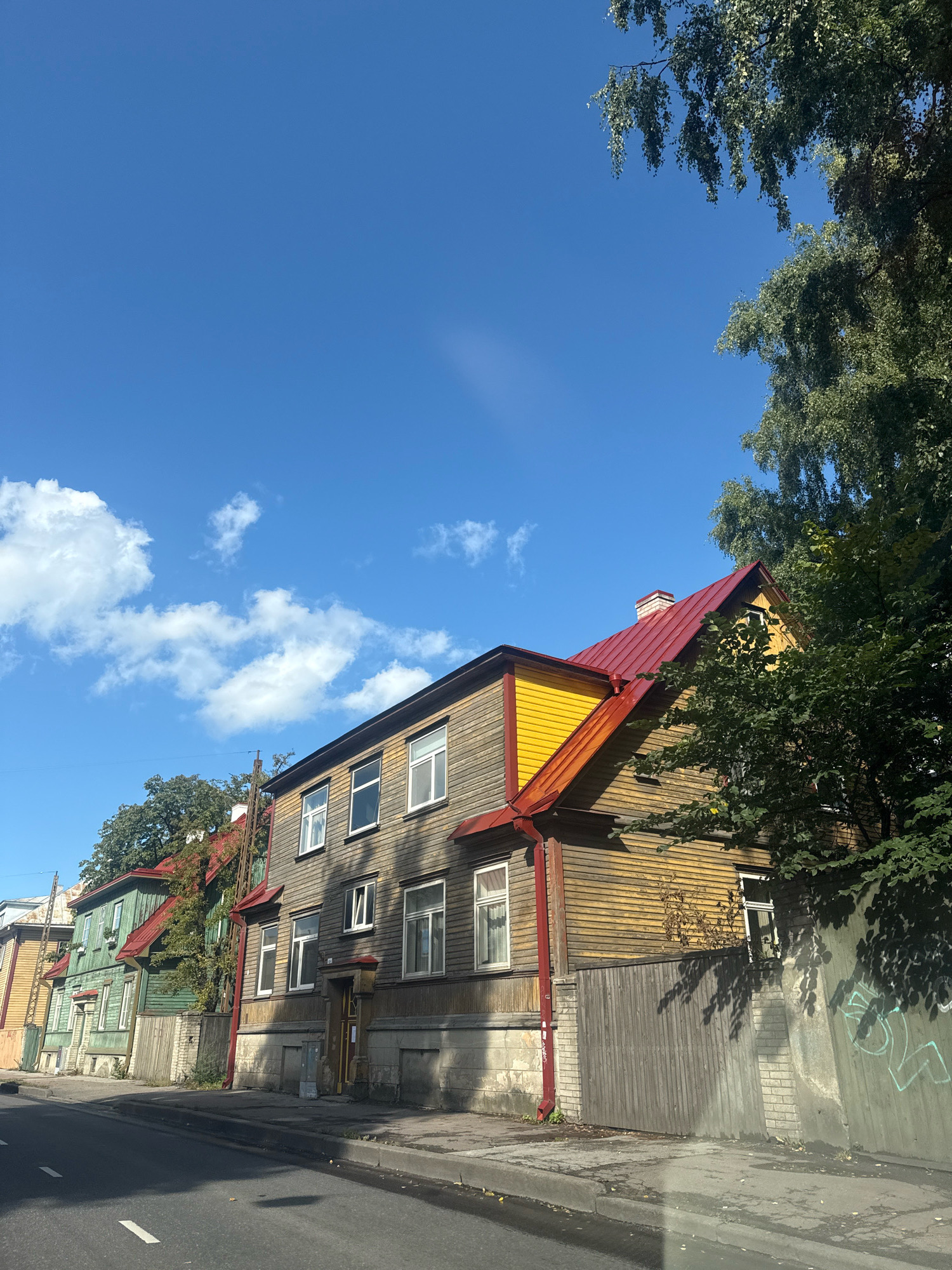 The image depicts a row of wooden houses with red roofs and yellow trim, located in Tallinn, Estonia. The houses appear to be old, with weathered wooden siding and traditional window designs. A paved street runs in front of the houses, with a sidewalk and a low wooden fence. Trees with green foliage partially obscure the houses, casting shadows on the scene. The sky is a clear blue with a few white clouds. The image is a medium-angle shot, taken from the street, capturing the facades of the houses and the surrounding environment. There are no people or animals visible in the image.
