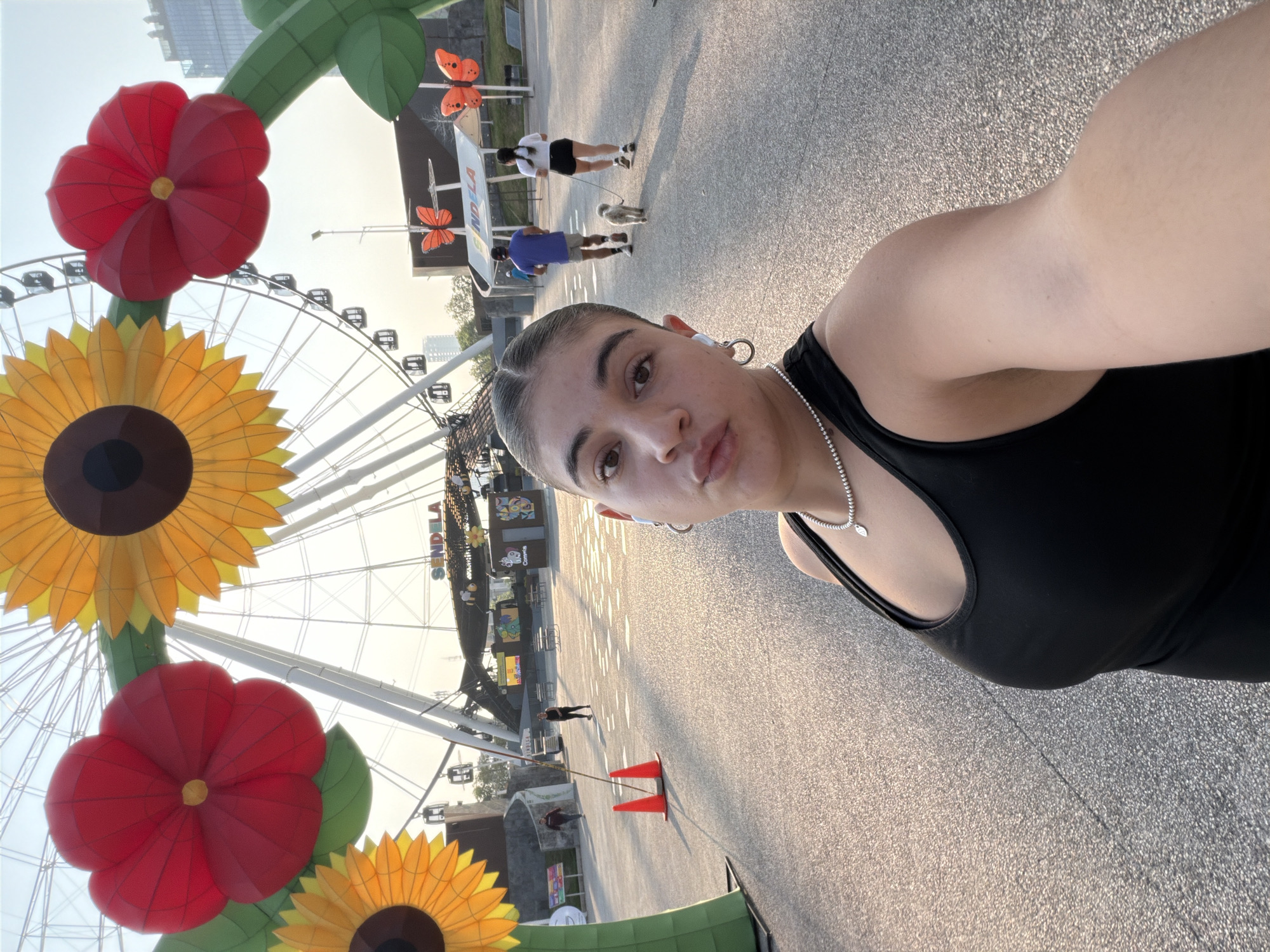 A person with light skin and braided dark hair, wearing a black tank top, silver necklace, and hoop earrings, takes a selfie on an overcast day. A large Ferris wheel and colorful flower-shaped decorations are visible in the background. Other people walk nearby. Orange traffic cones are visible in the distance. A sign with butterflies and the word "SENDELA" is also in the background. The location is Puebla, Mexico.