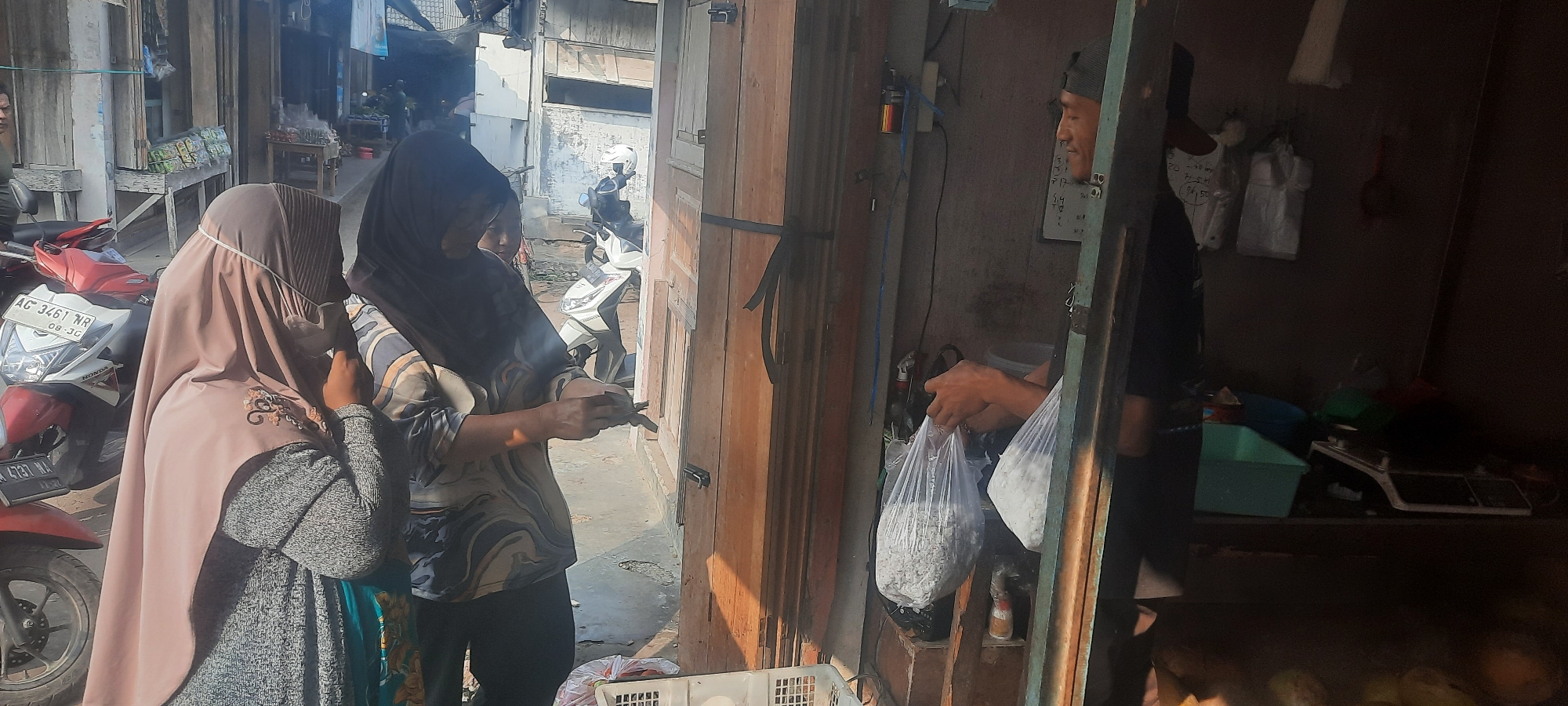 This image depicts an outdoor scene in Selopuro, Indonesia, likely a market or street vendor area. Two women, dressed in hijabs and modest clothing, are interacting with a man who is holding two plastic bags filled with white, granular material. The woman on the left wears a light pink hijab and a patterned dress, while the woman on the right wears a dark hijab and a patterned shirt. The man, wearing a dark t-shirt and a cap, appears to be handing over the bags.
In the background, a white scooter with a license plate is visible, along with some stalls and structures made of wood and what appears to be corrugated metal. The ground is paved and somewhat uneven, with some debris. Sunlight suggests it is daytime. The overall impression is a candid snapshot of a commercial transaction or interaction in a local Indonesian setting.