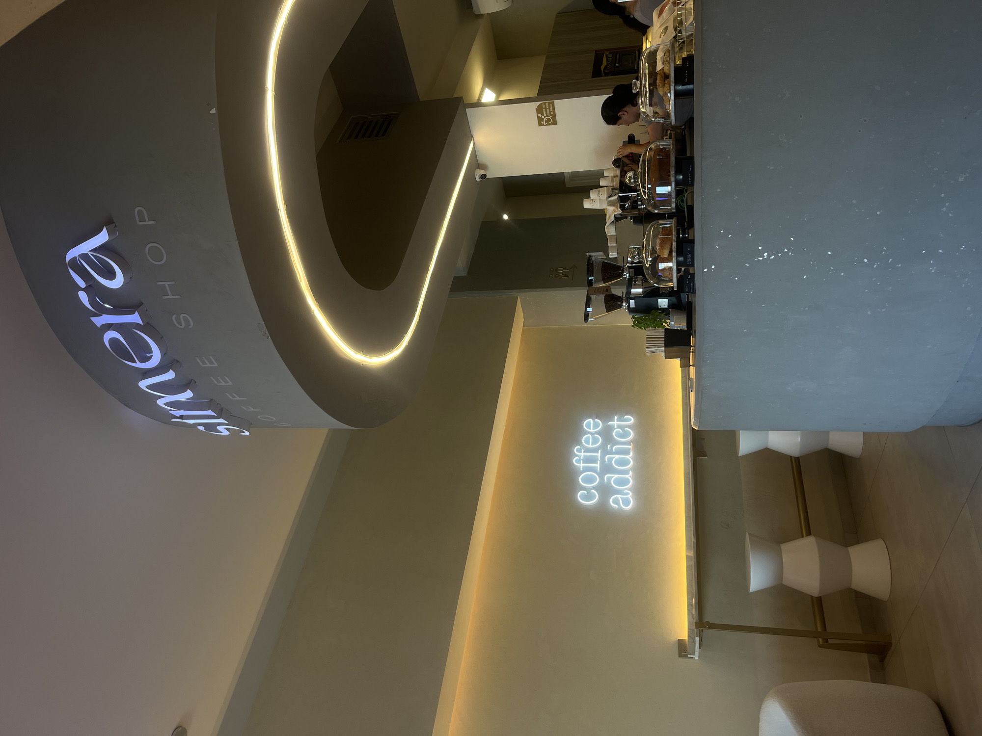 This image depicts the interior of a modern coffee shop, likely in San Bernardino Tlaxcalancingo, Mexico. The setting is indoors, illuminated primarily by artificial light, suggesting either nighttime or a deliberately ambient atmosphere.
The architecture features a prominent curved ceiling or wall section, accentuated by a warm, glowing strip light that follows its contour. On this curved surface, a luminous sign displays "Sumera" in a distinctive script font, with "COFFEE SHOP" printed in capital letters directly beneath it.
In the midground, a long, minimalist counter stretches across the frame. It is equipped with professional coffee machines, grinders, and several clear glass display cases containing pastries or other baked goods. Behind the counter, a staff member, presumably a barista, is seen attending to the equipment, looking down at their work. A second person's head is partially visible further down the counter.
A light-colored wall on the right side of the image features a bright, cool white neon sign that reads "coffee addict" in a script typeface. Below this, a seating area is implied by several modern, white, hourglass-shaped stools positioned along a counter with a metallic gold base. The overall aesthetic is clean, contemporary, and inviting, characterized by neutral color tones (light gray, beige, off-white) and thoughtful lighting design. A small security camera is also visible on a wall near the counter.