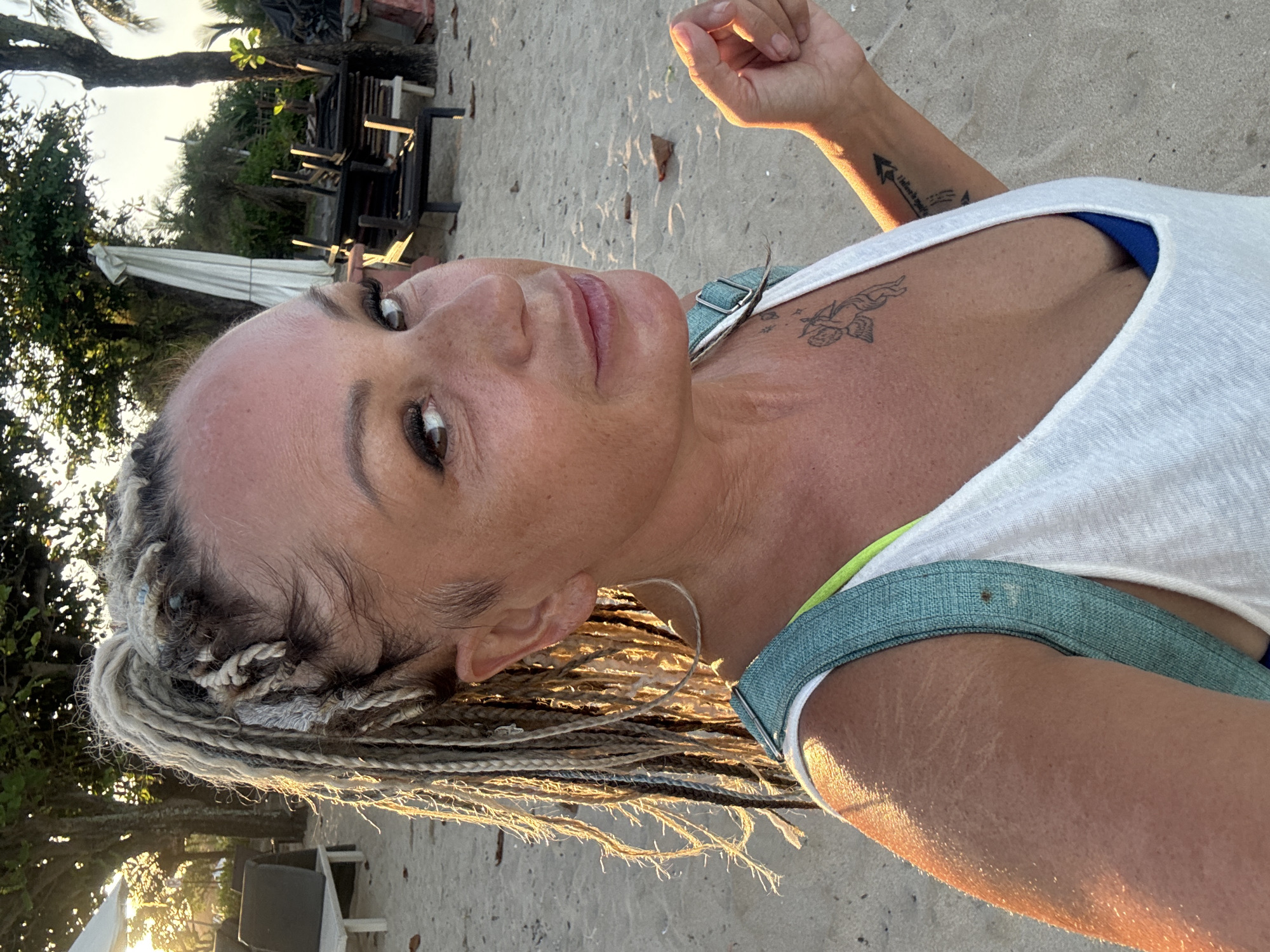 A woman with blonde braids is posing for a selfie on a beach. She is wearing a white tank top, revealing a blue swimsuit underneath. Tattoos are visible on her arm and chest. The background shows a sandy beach with trees, beach furniture, and what appears to be a white umbrella. The light suggests it might be early morning or late afternoon. Given the location, it is likely this beach is in Jimbaran, Indonesia.