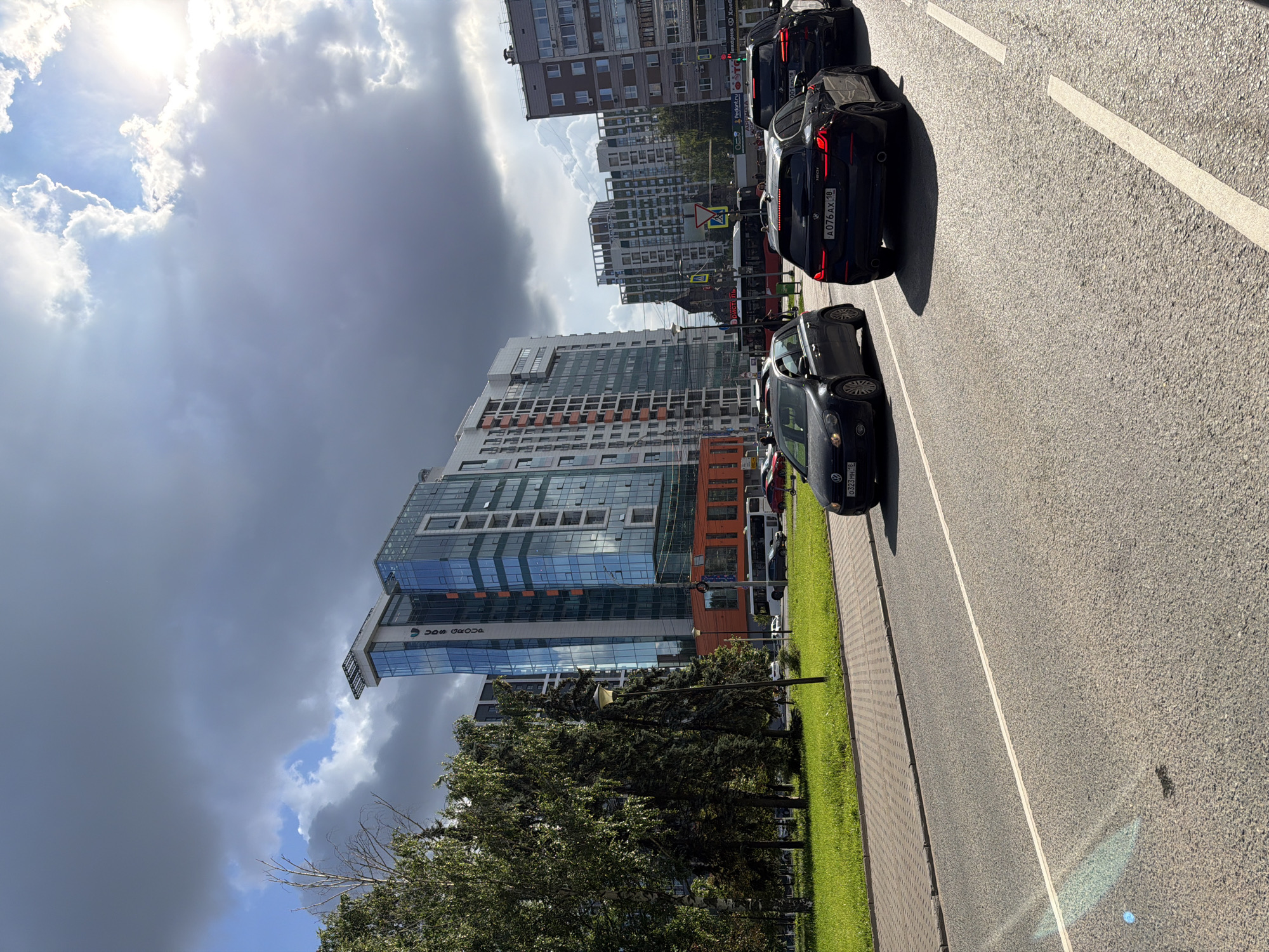The image depicts a street scene in Izhevsk, Russia, under a partly cloudy sky with the sun shining through. In the foreground, there are three cars on the road, a dark grey hatchback on the left, a black BMW wagon in the center, and another black car partially visible on the right. The BMW has the license plate "A076AX 18". The road is paved and marked with white lane lines. To the left of the road, there is a strip of green grass followed by a sidewalk paved with interlocking bricks. Beyond the sidewalk, there are several trees, including birch trees and evergreen trees.
In the background, a modern, multi-story building with a glass facade and orange accents stands prominently. This building appears to be an office building or residential complex. Further in the background, other multi-story buildings are visible, indicating an urban environment. A bus is also seen in the distance, suggesting public transportation is present. The overall scene is one of urban activity on a bright day.