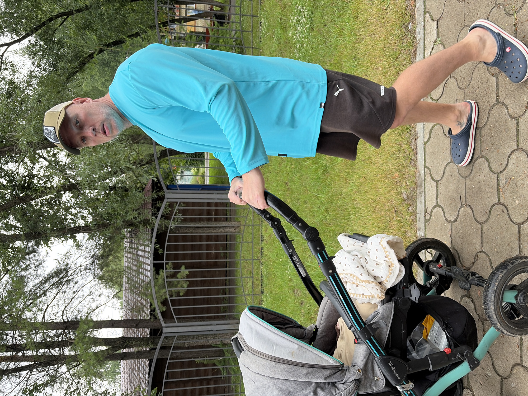 The image shows a man pushing a baby stroller on a paved path. He is wearing a light blue long-sleeved shirt, dark shorts, a beige baseball cap, and dark blue Crocs. The man is looking towards the camera with a neutral expression. The stroller is gray and black, with a light-colored blanket inside. In the background, there are trees, grass, and a metal fence. The scene appears to be outdoors, possibly a park or residential area. The lighting suggests it is daytime. The orientation of the image is vertical, with the subjects positioned in the lower half of the frame. The context provided indicates the location is Chaikovsky, Russia. There is some text visible on the stroller's handle, but it is too small to read clearly.