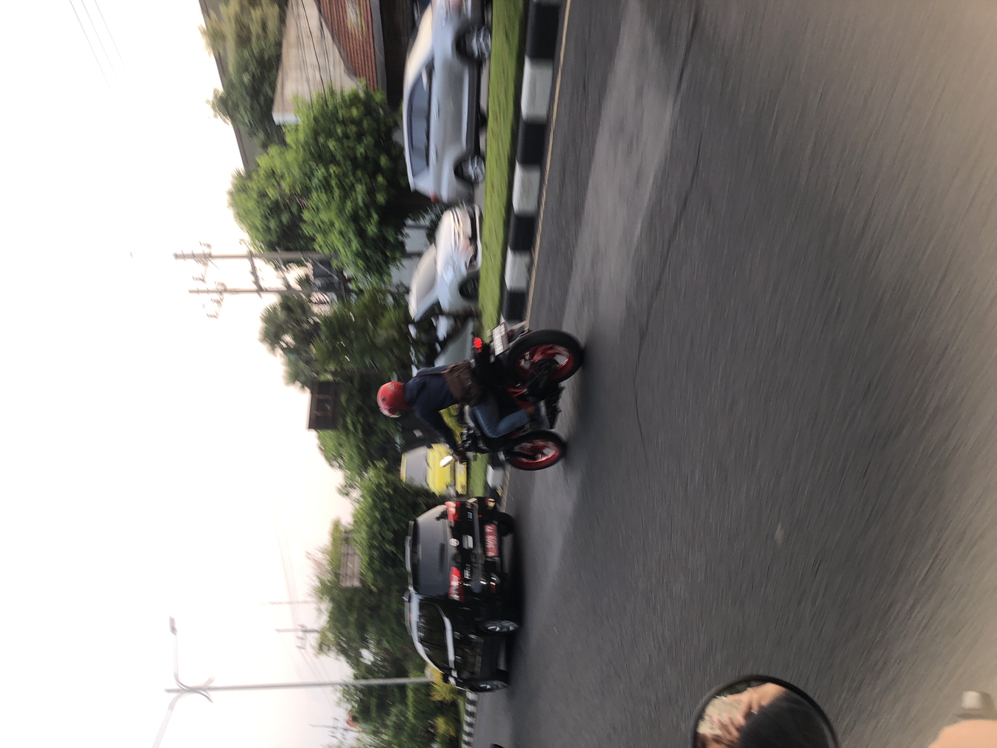 The image depicts a street scene in Klurak, Indonesia, captured during daytime with a slight motion blur. A motorcycle with a rider wearing a red helmet and jeans is moving from left to right across the frame. The motorcycle has red accents on its wheels. To the left of the motorcycle, a black SUV is also in motion, moving in the same direction. Further to the left, a street lamp is visible against a bright sky. In the background, there are trees and buildings, and other vehicles, including a yellow vehicle, are present on the road. The road is paved, and a grass median with black and white borders separates the lanes of traffic. The perspective suggests the photo was taken from another moving vehicle, possibly a motorcycle, as indicated by a partial view of a side mirror in the lower left corner reflecting a person's hand and arm.