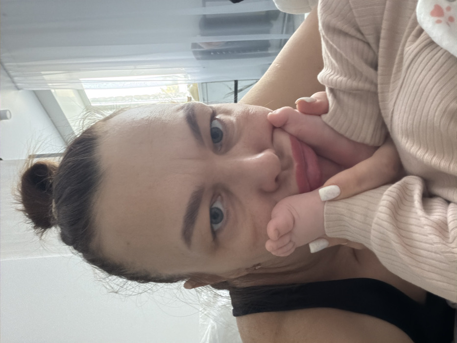 A woman with brown hair pulled back smiles at the camera. She has light skin and blue eyes. She is holding a baby, whose foot is covering part of her mouth. The baby is wearing a light pink sweater. The woman is wearing a black tank top. The background is a white wall and a window with blinds.