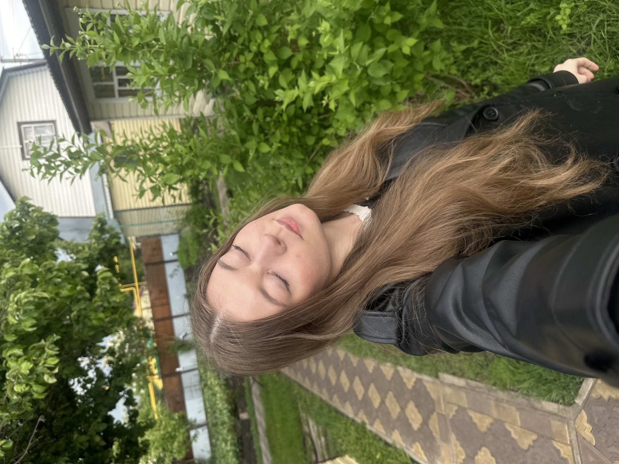 A medium shot of a person with long brown hair, closed eyes, and wearing a black leather jacket. They are standing outside on a paved walkway next to a grassy area and green bushes. In the background, there are residential buildings with light-colored siding. It is likely that the location is Коноваловка, Russia.