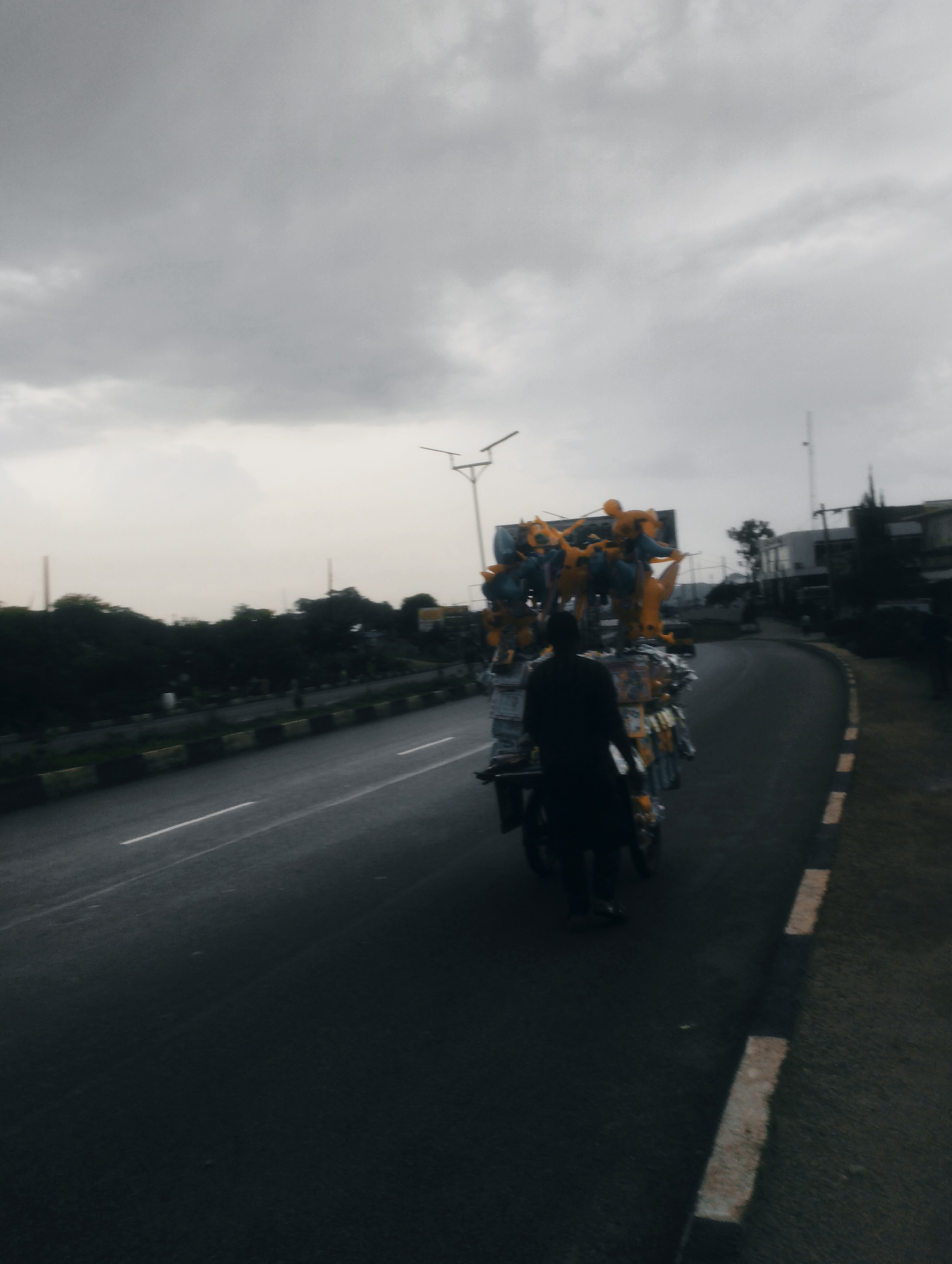 A person is riding a motorcycle loaded with yellow and blue items on a wet road, likely in Jos, Nigeria. The motorcycle is being driven on the right side of the road, with a lane to its left. The sky is overcast and gray. Trees and buildings are visible in the background. The road has a distinct white and black curb. The scene appears to be an urban environment.