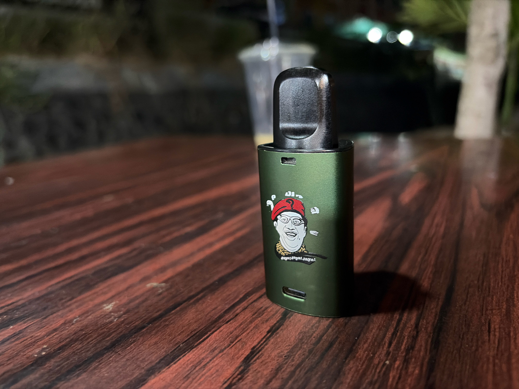 A close-up shot focuses on a green vape pen with a cartoon sticker of a man wearing a red beret and glasses, and the text "jaya! jaya! jaya!". The vape pen is resting on a dark, reddish-brown wooden table with visible grain. In the blurred background, a clear plastic cup with a straw is visible, along with greenery and out-of-focus lights, suggesting an outdoor setting at night. The image is likely taken in Tanjung Pinang, Indonesia, possibly at an outdoor dining area or cafe.