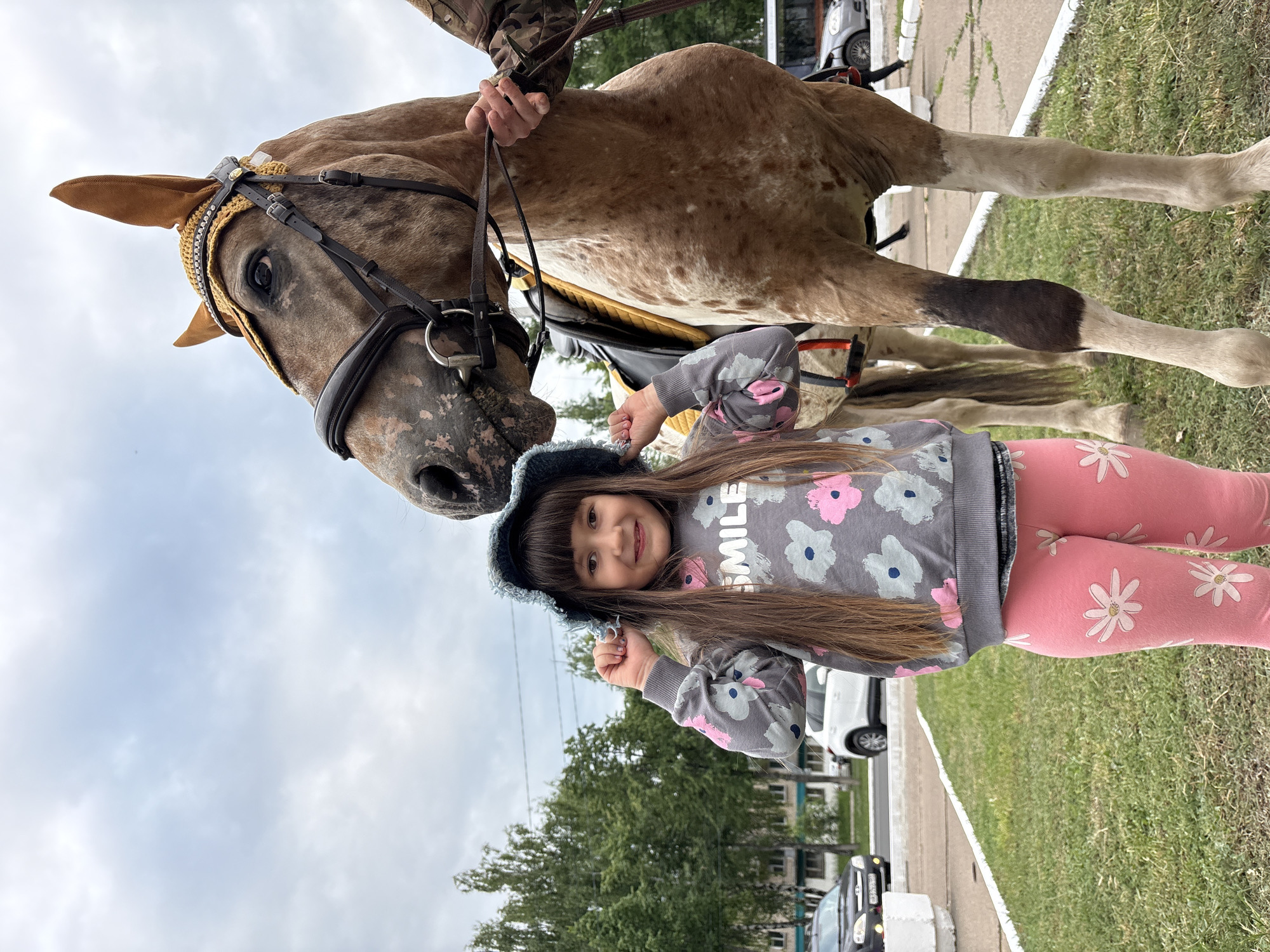 A young girl stands next to a horse in what appears to be a park or grassy area within a city. The girl, wearing a gray sweatshirt with floral patterns and pink leggings, is smiling and holding her blue hat. The horse, a brown and white spotted breed, is wearing a decorative bridle and ear covering. In the background, there are trees, buildings, and cars, suggesting an urban environment. The sky is overcast.