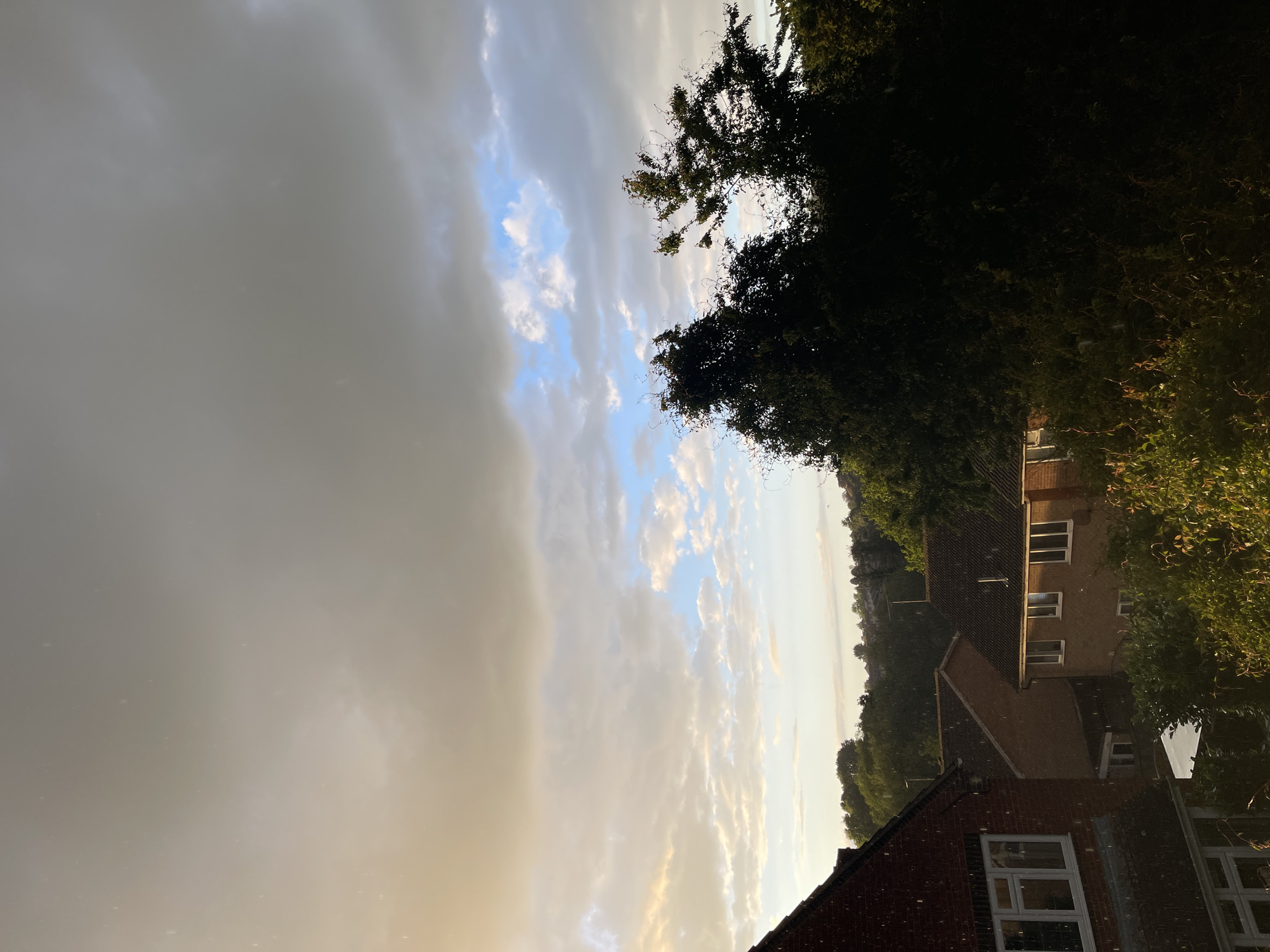 The image shows a residential area in Maidstone, United Kingdom, taken outdoors under a cloudy sky. To the left, heavy grey clouds dominate the view, while a strip of blue sky with white clouds is visible in the center. In the foreground, the roofs of brick houses are visible, with white-framed windows. Trees and foliage fill the right side of the image, partially obscuring some of the houses. The photograph is slightly tilted counter-clockwise. There are no people visible.