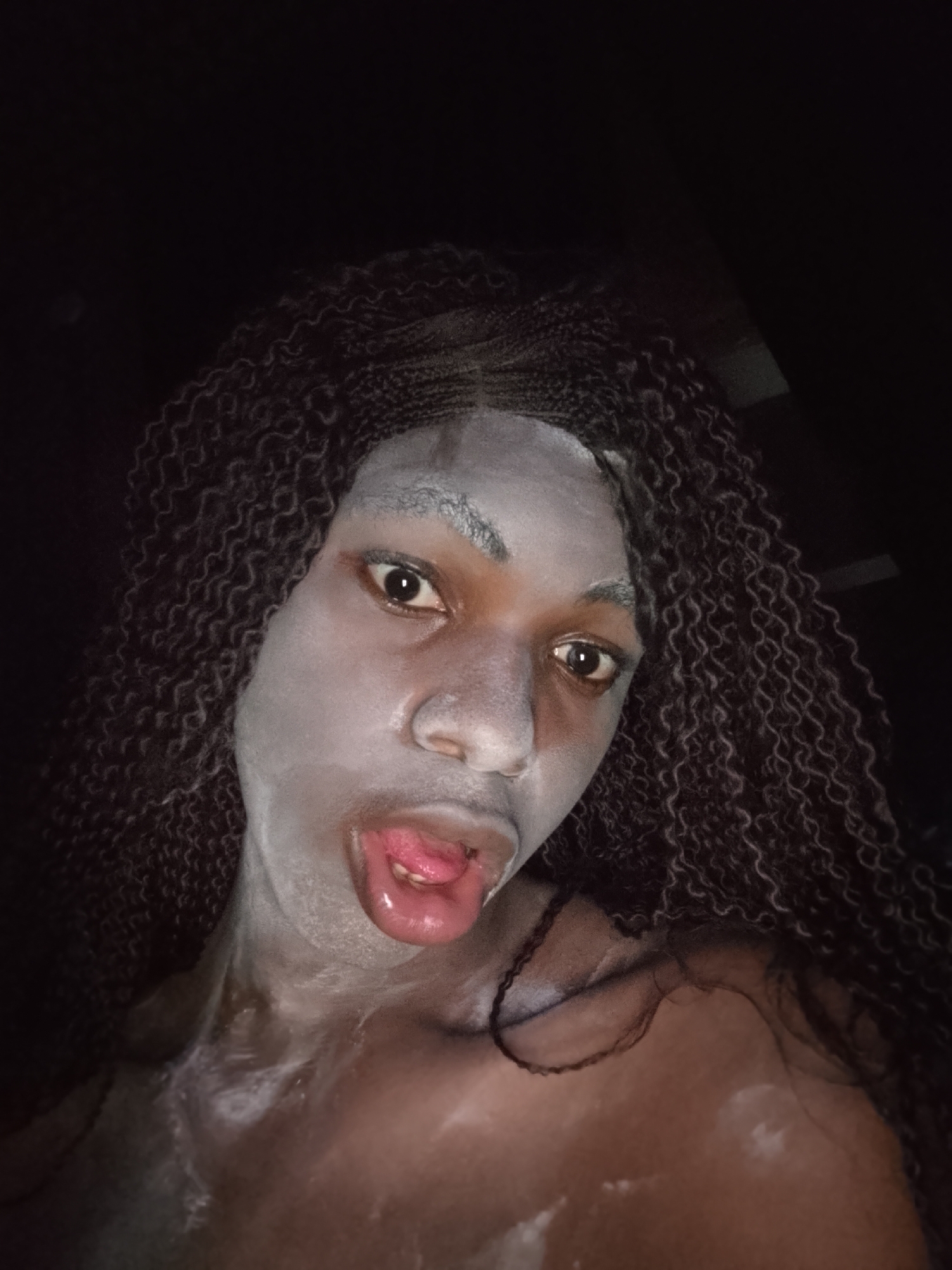 A close-up, dimly lit portrait shows an individual with dark skin, looking directly at the camera. Their face, neck, and upper chest are partially covered in a light grey or white, mask-like substance, suggesting a beauty treatment or artistic makeup. Long, dark, curly braided hair frames the face and extends over the shoulders. The individual's eyes are wide open, and their mouth is open with the tongue slightly protruding, revealing its pink surface and the inner part of the lips, which appear wet or glossy. The dark background offers no discernible features, placing full emphasis on the person. The overall impression is one of a playful or expressive moment captured in low light. No text is visible.