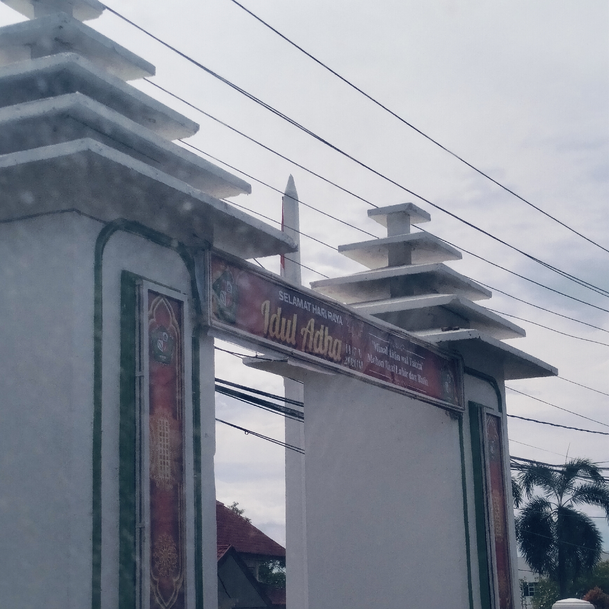 The image displays an ornate white archway or entrance gate, situated outdoors, likely in Lampeudeu Baroh, Indonesia. The archway features green trim along its edges and decorative panels with intricate patterns on its pillars. Each pillar is topped with multi-tiered, stepped architectural elements.
A red banner stretches across the top of the archway, bearing text in gold and white. The banner reads: "SELAMAT HARI RAYA Idul Adha," indicating the celebration of Eid al-Adha. Below "Idul Adha," the text "10/V H" and "2023 M" are visible, likely denoting the date of the holiday. To the right, additional greetings state "Minal 'Aidin wal Faizin" and "Mohon Maaf Lahir dan Batin," which are common Indonesian Eid greetings for seeking and granting forgiveness.
In the background, a tall, white pointed structure, resembling a minaret, rises against an overcast, cloudy sky. Several black electrical wires crisscross the upper portion of the frame. Through the archway on the left, a building with a reddish-brown tiled roof is partially visible. On the right, a large palm tree and other green foliage add to the natural elements of the scene. The image appears to be taken during the daytime under a soft, diffuse light from the cloudy sky. No people are present.