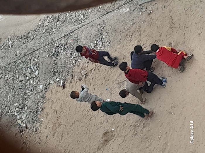 An elevated view captures seven young males, identified as children or pre-teens, on a sloping sandy and dirt terrain in Khan Yunis, Palestinian Territory. The ground is predominantly light brown soil, disturbed in various areas. To the upper right, the terrain transitions into a steep incline covered densely with grey rubble, rocks, and debris; a dark, thin cable or wire runs diagonally across this rubble.
The children are positioned across the sandy area, engaged in a downward-focused activity. From left to right:
1. A child in a light grey tracksuit stands facing down and slightly right.
2. A child in a dark green tracksuit stands facing forward, barefoot.
3. A child in a light brown/tan top and dark pants is crouched low, hands on the ground.
4. A child in a red top with dark blue accents and dark pants stands, leaning forward and looking down.
5. A child in a red hoodie and dark pants is leaning forward with hands on the ground, wearing blue and white sneakers.
6. A child in a dark blue top and dark pants is on hands and knees, facing left.
7. A child in a red and yellow tracksuit is on hands and knees, facing the bottom right, wearing dark flip-flops.
The children's postures indicate individual focus on the immediate ground directly in front of them, suggesting an activity or exploration of the surface. No explicit objects or tools are visible in their hands. The ambient light is uniform, with no strong shadows. A "Galaxy A14" watermark is visible in the lower left corner.