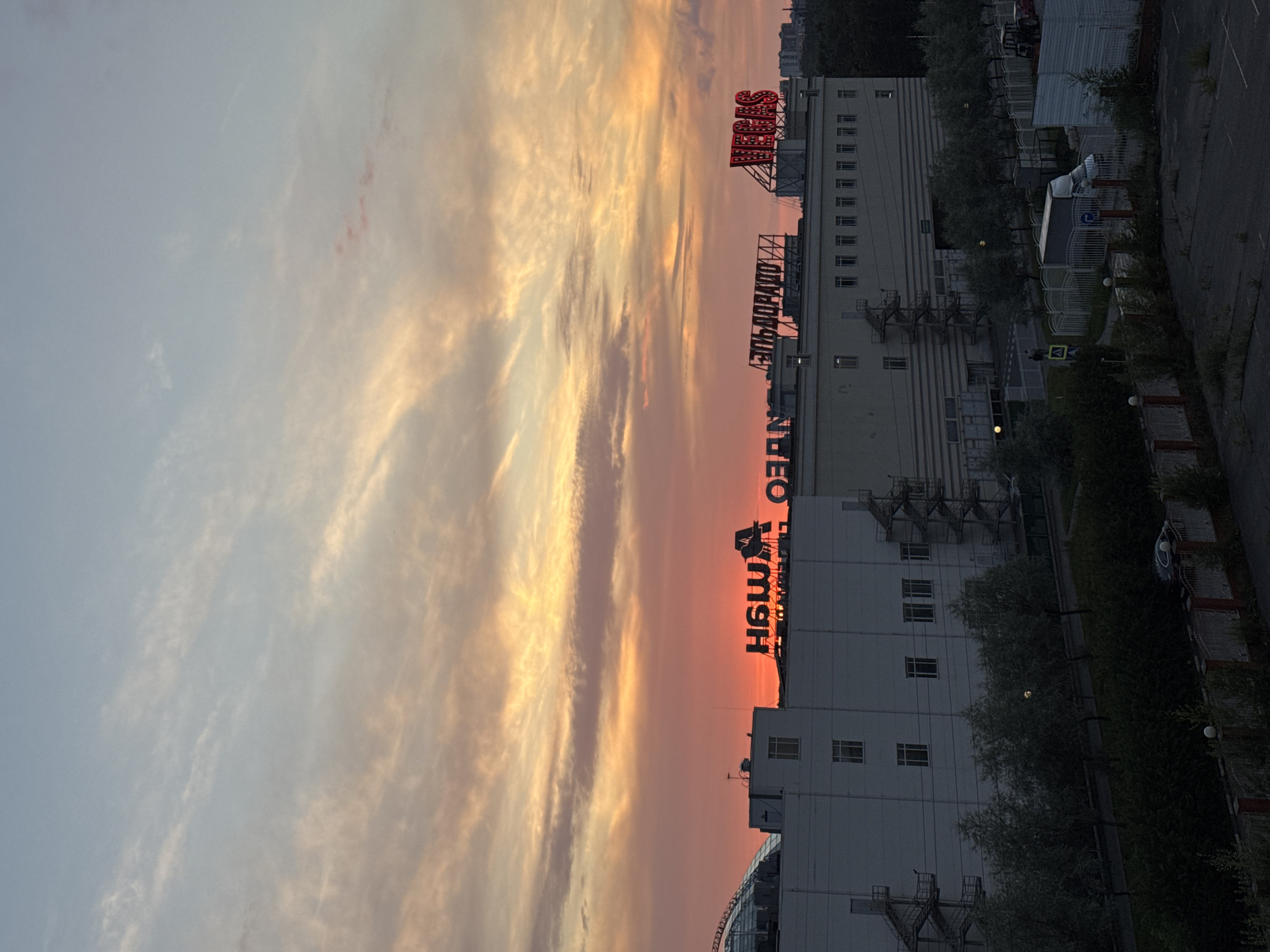 The image captures a sunset over a commercial building in Sovkhoza imeni Lenina, Russia. The sky is filled with hues of orange, pink, and yellow due to the setting sun, with wispy clouds scattered across the expanse. Dominating the mid-ground is a large, white, multi-story building with a modern glass-domed section on the left. Several signs are visible on the rooftop of the building. To the right, a red neon sign that reads "VEGAS" is illuminated. To the left of "VEGAS" is another sign, possibly in Russian, with Cyrillic lettering that appears to read "ЭЛЬДОРАДО". Further left, another sign with Cyrillic lettering, "НАШ ДОМ", is also visible. In front of the building, rows of trees with sparse foliage line a paved area. A white truck is parked behind a fence on the right side of the image. The foreground shows a dark, paved area with some debris. The overall scene suggests an urban or suburban commercial area during the evening.